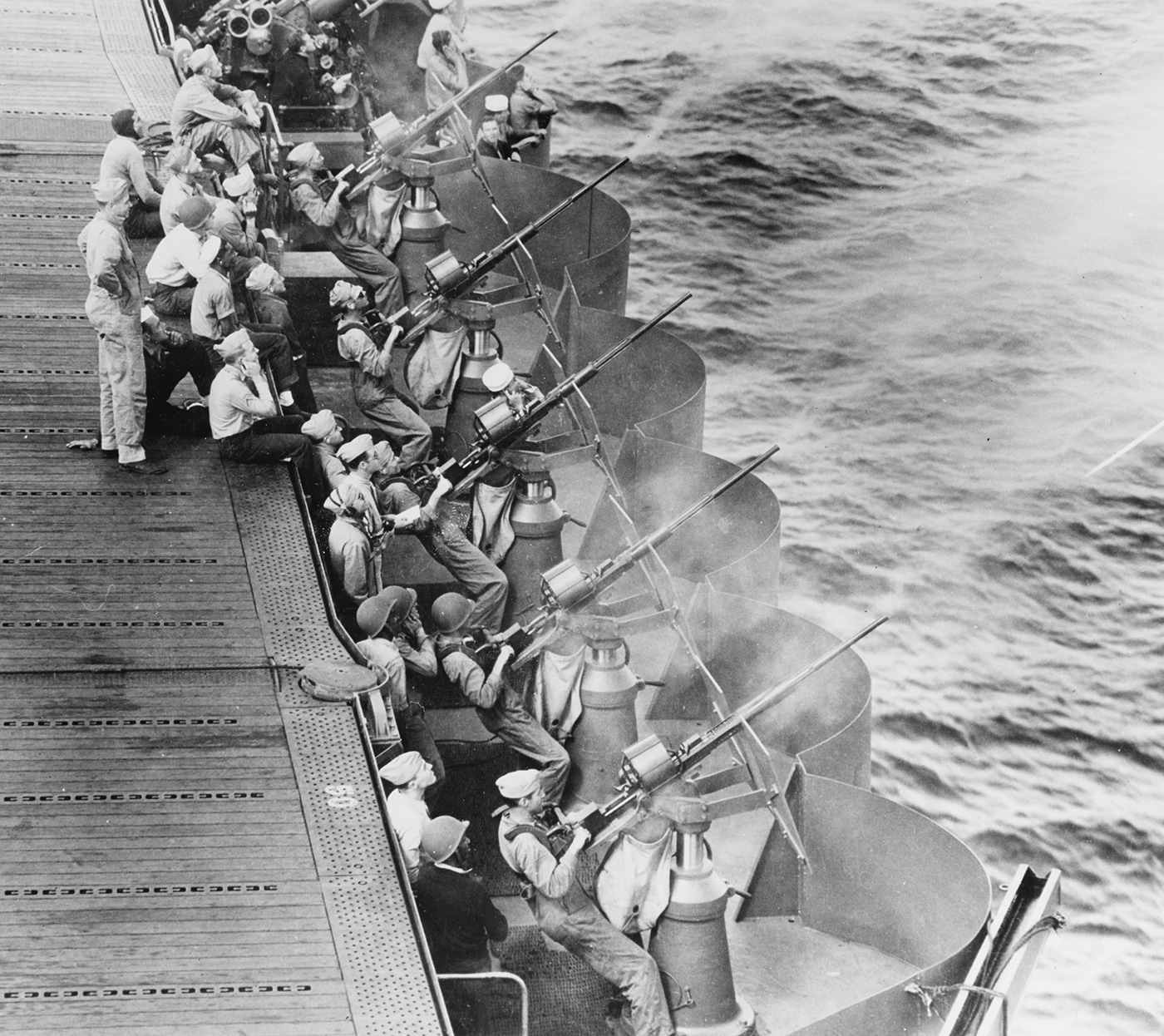 This photo shows sailors aboard USS Enterprise (CV-6) manning Oerlikon 20mm anti-aircraft cannons as they prepare to defend against Japanese air attacks during World War II. The gunners are at their stations along the edge of the flight deck, hands on the controls and eyes trained on the horizon for incoming enemy planes. Each cannon is mounted on a swivel, with ammunition drums in place and protective shields surrounding the position. The sailors wear helmets, life vests, and other safety gear, fully alert to the possibility of imminent attack. In the background, the flight deck and other defensive gun positions can be seen, creating a layered network of protection around the carrier. The image highlights the crucial role of these small but powerful cannons in repelling dive bombers, torpedo planes, and kamikaze threats. It is a clear example of the daily vigilance and teamwork required to keep USS Enterprise safe in hostile waters.