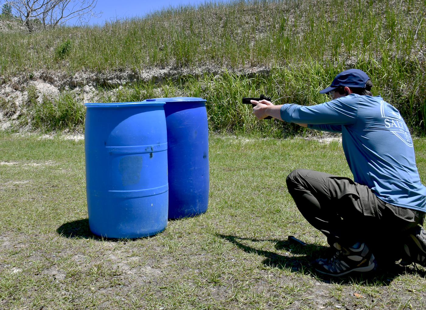 A man is shown firing his pistol around the side of a plastic drum used as cover on an outdoor range, keeping his body low and only exposing a minimal profile. The muzzle is angled carefully to reach the paper targets while most of his torso remains protected behind the drum. This image demonstrates a key skill taught in two self-defense shooting drills — how to use cover to reduce exposure while continuing to deliver accurate shots. The scene includes visible target frames and a clear shooting lane, emphasizing a controlled training environment. The shooter’s posture highlights muzzle discipline, sight alignment, and deliberate trigger control. This picture is useful for illustrating tactical movement, cover use, and practical defensive handgun techniques.