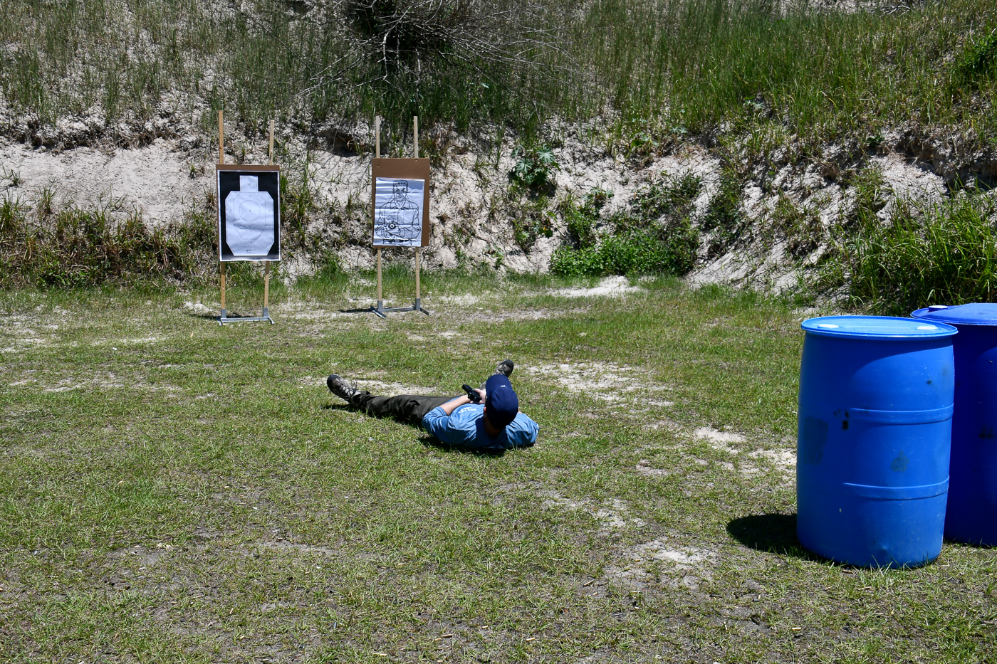 On the range, a shooter lies supine on the ground at the start of a defensive drill. His pistol is safely holstered as he positions himself for the first stage of training. This drill simulates being knocked down by an attacker and forces the shooter to recover while under threat. Targets are placed a few yards ahead, and training equipment such as plastic drums are set up to simulate cover. The drill highlights how two self-defense shooting drills teach shooters to handle disadvantaged positions. Practicing from the ground ensures familiarity with movement, safe gun handling, and tactical accuracy.