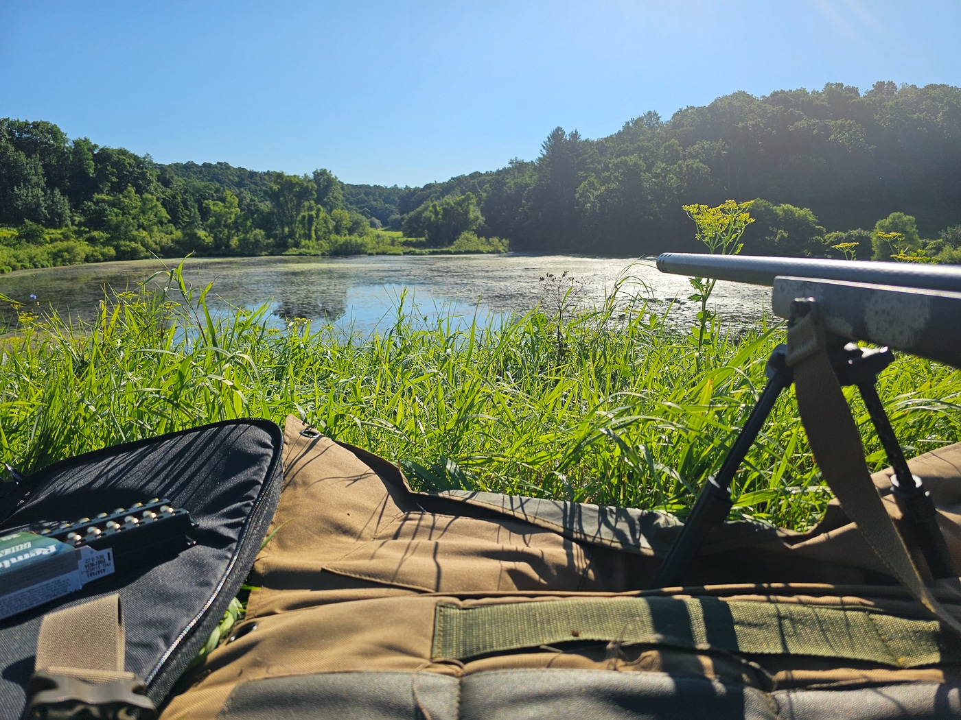 A shooter engages distant steel targets during the Field Course Test of the Vortex Optics Hunter Marksmanship Course. The outdoor range stretches across open terrain with mixed grass and slight elevation changes, offering distances from 50 to 500 yards. The participant kneels behind a rifle supported on a backpack, with an instructor timing the stage nearby. Wind flags ripple in the background while other shooters observe their peers’ performance. This scene illustrates how the course simulates real hunting conditions where range estimation, position building, and shot timing are critical. The image captures the culmination of two days of training  —  applying classroom knowledge and indoor range data in a practical, outdoor setting.