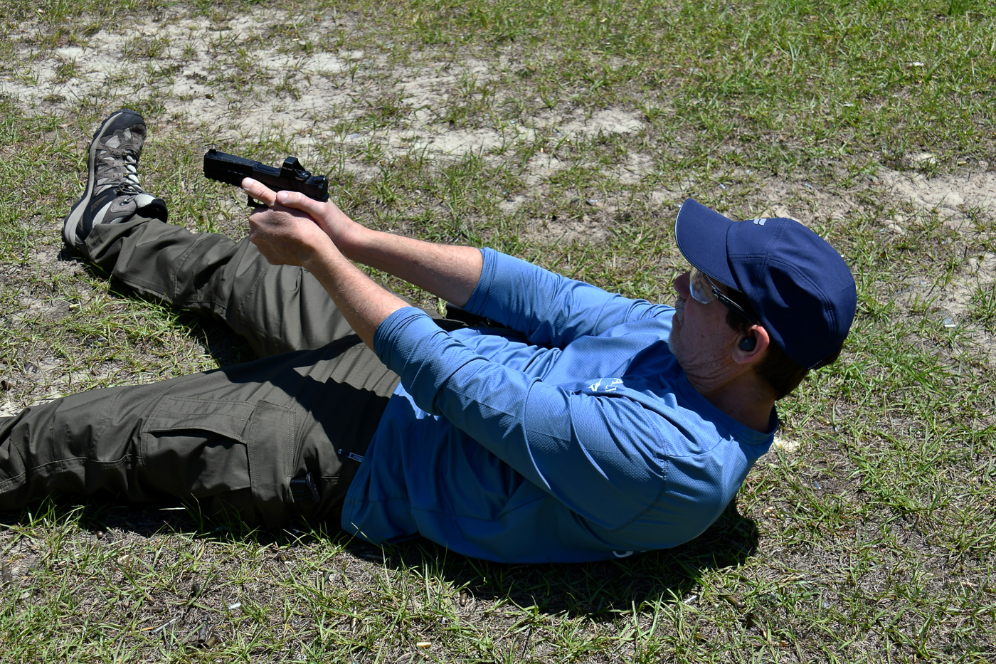 A shooter is pictured mid-drill on an outdoor range, lying on his back with his upper torso raised as he fires at paper targets. His arms are extended in a two-handed grip, showing proper muzzle control and sight alignment. This drill simulates defending oneself immediately after being knocked down in an attack. Targets are positioned close by, emphasizing speed and accuracy under stress. The training is part of two self-defense shooting drills designed to prepare shooters for realistic survival conditions. Practicing from the ground ensures familiarity with unconventional firing positions, recovery movements, and safe gun handling.