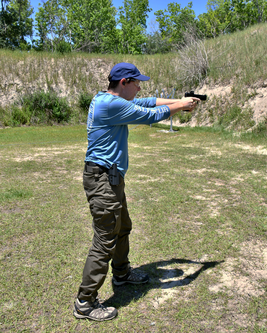 A man is shown on an outdoor range firing his handgun from an open position without nearby hard cover, demonstrating how to respond when shelter isn’t available. The image highlights rapid, accurate shooting and controlled recoil management under simulated stress. This scene relates to two self-defense shooting drills that teach both seeking cover and, when necessary, applying suppressive accurate fire while moving. Visible targets and clear lanes emphasize a controlled training environment where exposure and movement are practiced safely. The photograph underscores the balance between speed and precision when fighting from the open. It also reinforces basic safety: muzzle awareness, proper stance, and eye protection.