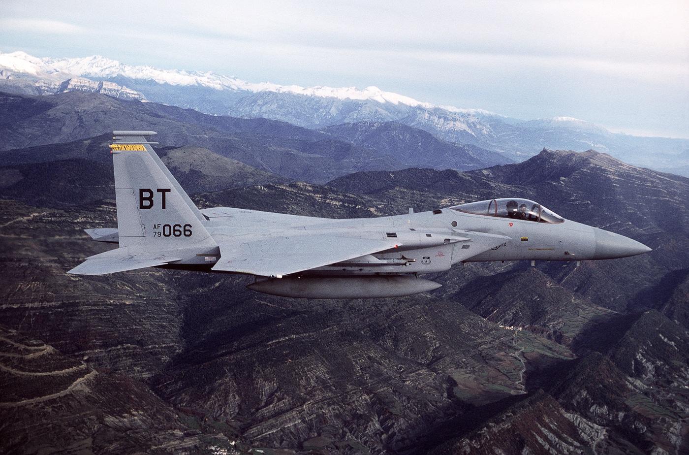 A right-side, air-to-air photograph shows a U.S. Air Force F-15C Eagle from the 53rd Tactical Fighter Squadron flying over Spain. The aircraft’s twin vertical stabilizers, swept wings, and gray camouflage pattern are clearly visible against the bright landscape below. The F-15C is a single-seat variant designed for air superiority missions, equipped with advanced radar and missile systems. This photo captures the fighter during a training deployment supporting NATO readiness and joint operations. The image emphasizes the power and agility of the McDonnell Douglas F-15 platform. The clear sky and sharp sunlight enhance the view of the aircraft’s aerodynamic form and operational markings.
