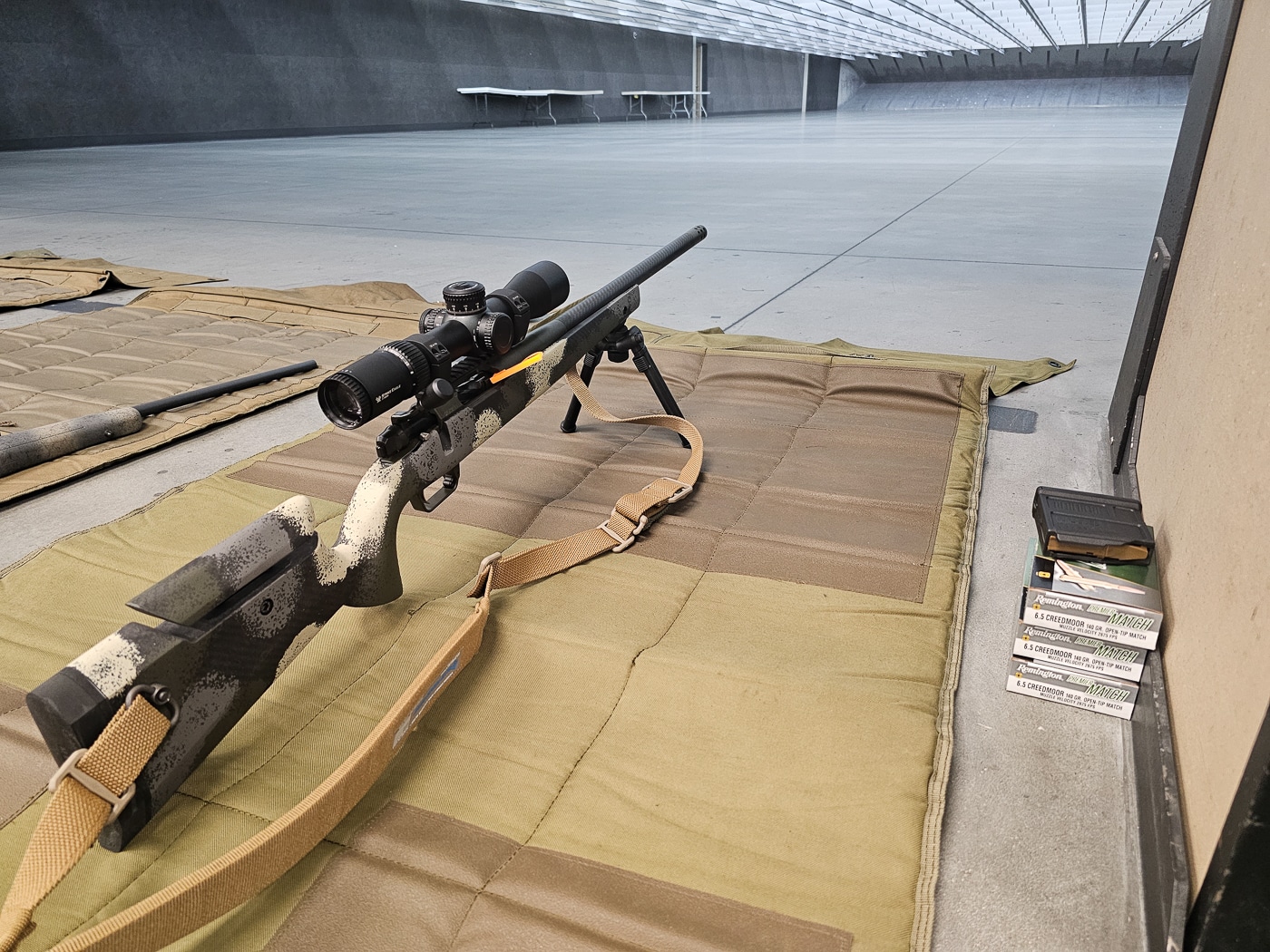 Indoor range scene showing a hunter sighting in a hunting rifle during the Vortex Optics Hunter Marksmanship Course. The rifle is supported on a bench with a bipod and the shooter wears ear and eye protection while peering through a magnified riflescope to make adjustments. Targets are set at a known distance downrange; brass and ammunition boxes lie nearby on the bench. An instructor stands slightly behind, observing and calling corrections as the student fires controlled groups to confirm or change the zero. The image emphasizes the process of turret adjustments, reticle interpretation, and confirming first-round hits before taking the same setup outside. This step is essential for accurate DOPE creation and ethical shot placement in the field.
