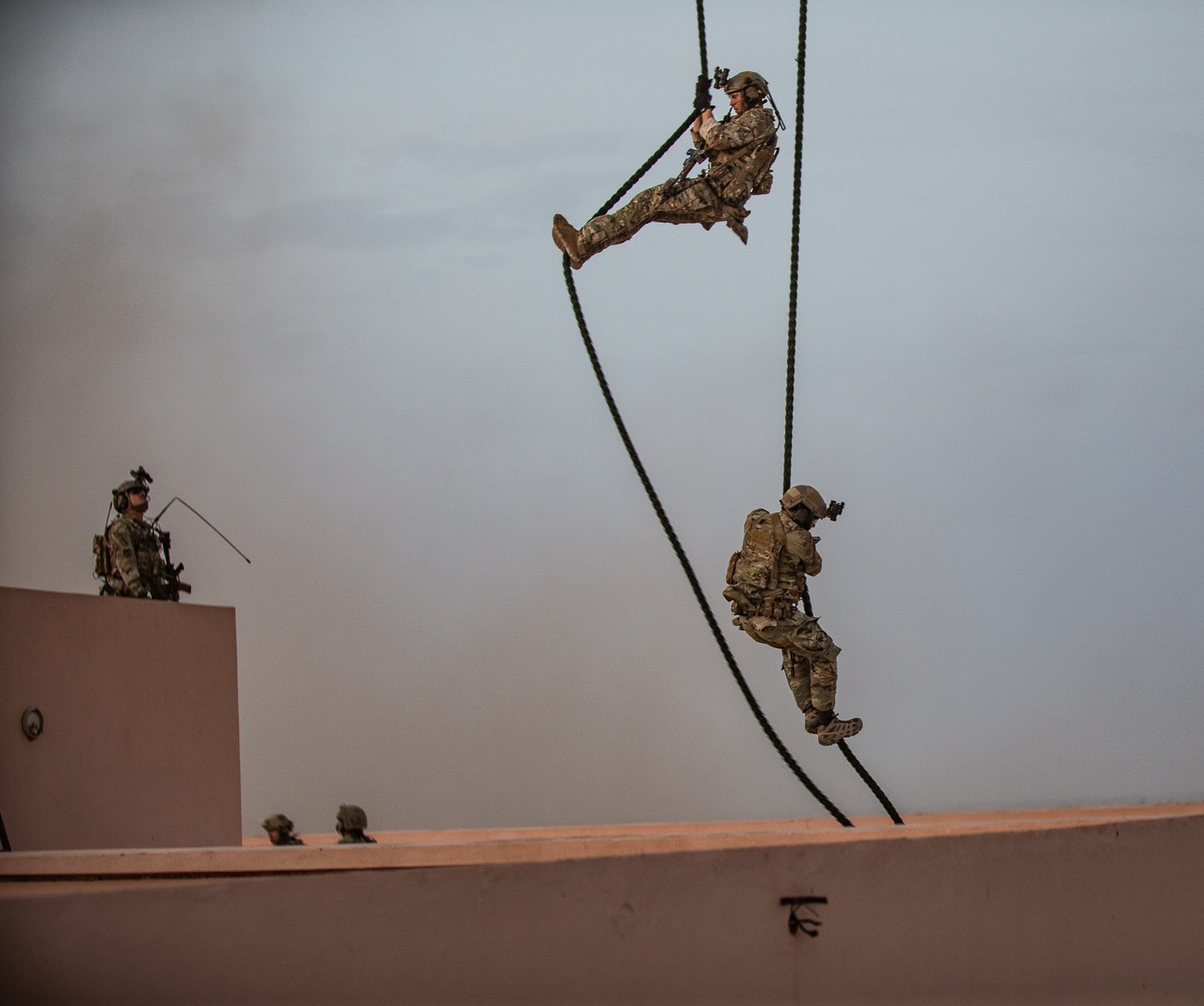 The image shows multiple U.S. Army soldiers from the 19th Special Forces Group fast roping from the rear or side doors of a 160th Special Operations Aviation Regiment (Airborne) MH-47 Chinook helicopter during African Lion 2021 in Morocco. The helicopter hovers low above rugged, dusty terrain with its rotors spinning, and thick ropes extended down to the ground. Soldiers, fully equipped with helmets, gloves, and tactical gear, are captured mid-descent, focused and moving quickly. The Moroccan environment is visible below, featuring arid, open ground and sparse vegetation, with clear skies overhead. This scene highlights the collaboration between U.S. special operations aviators and Special Forces in North Africa. The fast rope insertion technique allows for rapid deployment of personnel in locations where landing is not possible. The image demonstrates interoperability, operational readiness, and partnership as core aspects of African Lion.