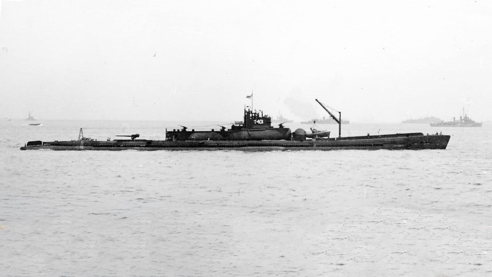 A black-and-white photo showing the Imperial Japanese Navy submarine I-401 on the surface after Japan’s surrender on September 13, 1945. The submarine’s long hull and large conning tower dominate the image. Visible on the aft deck is a Type 11 5.5-inch gun, used for surface defense. The I-401 was one of the I-400 class submarines, designed to carry and launch Aichi M6A Seiran floatplanes from a watertight hangar at the front of the vessel. This image captures the immense size and complex design of Japan’s underwater aircraft carriers as they were inspected by Allied forces after World War II.