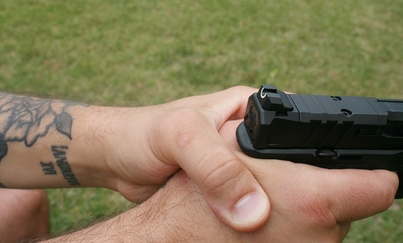 Image showing improper thumb positioning on a pistol grip with the thumb placed dangerously over the back of the grip frame where it will contact the moving slide. When the pistol is fired the slide travels rearward with significant force and any body part in its path will be struck causing painful injury. Correct grip technique positions both thumbs forward along the frame of the pistol keeping them clear of all moving parts and external controls. The dominant hand thumb should maintain contact with the frame on 1911-style pistols or be positioned high along the frame on other designs. Support hand thumb placement should be underneath the dominant thumb also oriented forward. This fundamental mistake demonstrates why shooters must understand how their pistol operates mechanically before establishing their grip and why proper fundamentals training is critical for safe shooting.