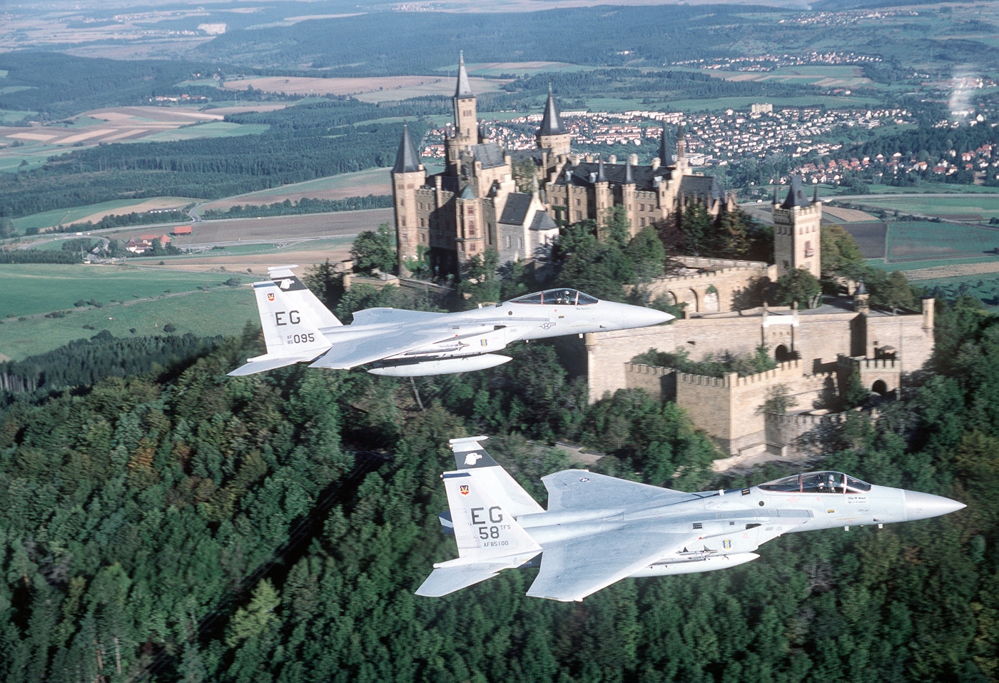 Two F-15C Eagle aircraft from the U.S. Air Force’s 58th Tactical Fighter Squadron fly in formation past Hohenzollern Castle, south of Stuttgart, West Germany. The air-to-air photo was taken during a Coronet Phaser deployment, part of NATO’s Cold War-era readiness exercises. The fighters are seen mid-flight, their gray camouflage contrasting against the green hills and historic architecture below. The Hohenzollern Castle, perched on a mountain ridge, provides a dramatic backdrop that emphasizes the blend of modern military technology and European history. Each F-15C features twin vertical stabilizers, swept wings, and missile pylons visible under the wings. The image captures both the strength of the U.S. Air Force and its cooperation with NATO allies in Western Europe.