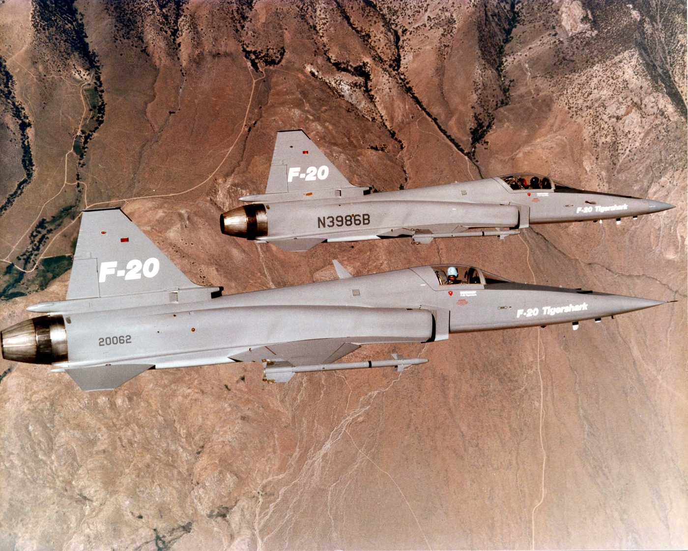 Two Northrop F-20 Tigershark fighter jets are seen flying side by side in a smooth formation at medium altitude. The aircraft maintain even spacing and level flight, showing precise aerodynamic control. Their light gray paint schemes and compact, single-engine designs are visible against a clear blue background. The photo was likely taken during a test or promotional flight to demonstrate stability and coordinated performance. Both aircraft feature the distinctive sharp nose and mid-mounted wings characteristic of the F-20. The sunlight reflects softly off their fuselages, emphasizing clean design lines. The scene captures the Tigershark’s balance of power and simplicity in flight.