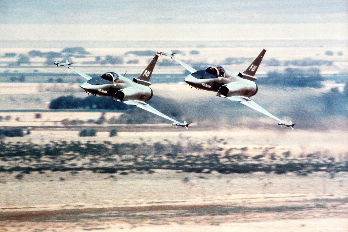Two Northrop F-20 Tigershark fighters are seen performing a high-speed, low-level pass during a demonstration flight. The aircraft fly in close formation just a few hundred feet above the ground, their gray paint schemes glinting in the sunlight. Heat distortion is visible behind the jets as their engines produce shimmering exhaust trails. The blurred landscape below conveys the immense speed of the maneuver. Northrop often showcased the Tigershark’s power and stability with low-altitude passes during public demonstrations. The aircraft maintain tight spacing, demonstrating precise control and aerodynamic balance. This photo captures the intensity and professionalism of the F-20 test and marketing flights.