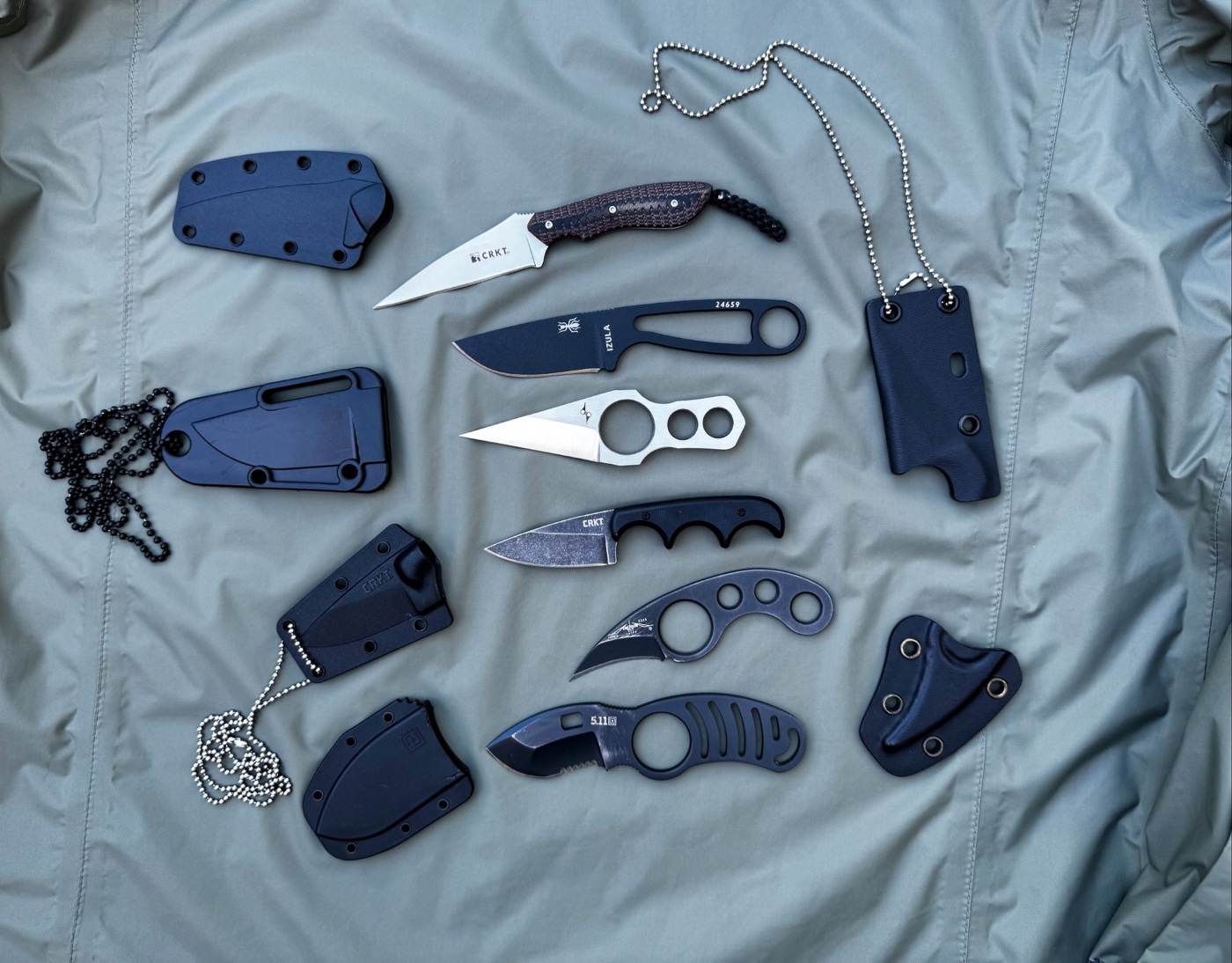 A selection of compact neck knives is laid out for comparison, each with a different style of handle featuring either finger grooves or integrated finger holes. Some knives have deep grooves along the handle scales to fit three fingers, while others include a forward finger ring for added retention. The knives vary in blade profile, from straight-edged Wharncliffe styles to more curved utility shapes. Each blade is paired with a molded sheath and short chain or lanyard suitable for neck carry. The arrangement emphasizes how ergonomic features can help smaller knives stay secure and comfortable in use. This ALT text is optimized for screen readers and includes terms like neck knife, finger grooves, finger hole, fixed blade, and sheath.