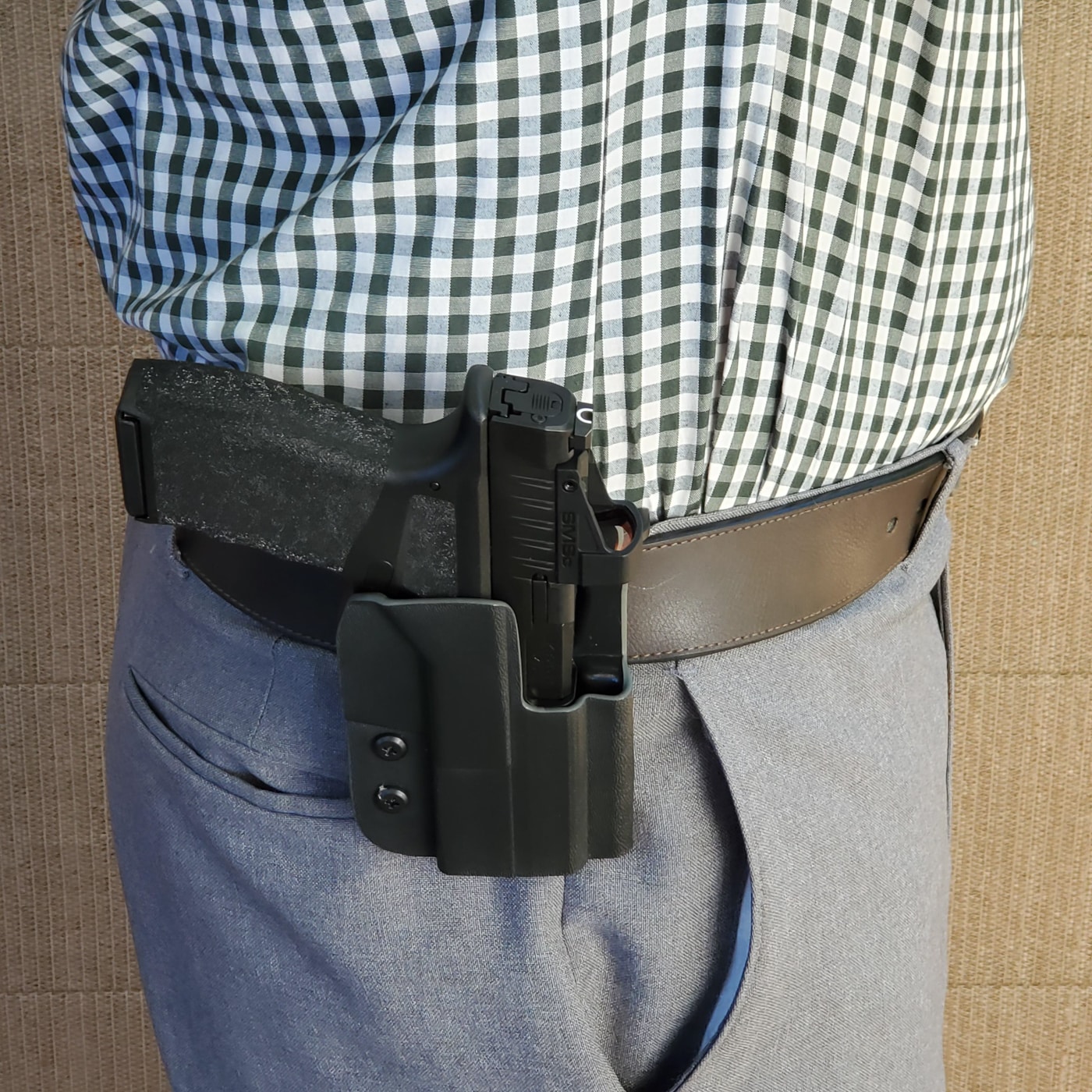 A person wearing a paddle holster mounted on their belt, demonstrating how the paddle secures inside the waistband while the holster body rests outside. The holster sits at the 3–4 o’clock position, a common placement for right-handed shooters. The paddle attachment grips firmly against the belt and waistband to prevent shifting during movement. This setup allows for fast on-and-off use, unlike traditional belt-loop holsters that require unthreading the belt. The pistol grip is visible for a quick draw while still positioned close to the body for concealment under a jacket. Keywords: paddle holster on belt, OWB holster carry position, concealed carry holster, waistband paddle attachment. The image highlights real-world use of this holster style.