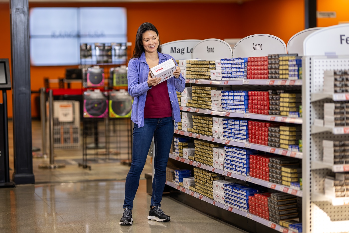 Photo of a woman standing in front of an ammunition display at Range USA, examining a box of cartridges before purchase. The shelves behind her are stocked with various calibers and brands, arranged neatly for easy browsing. The store is brightly lit and organized, with clear labels and visible Range USA branding. The customer appears focused and comfortable, representing the approachable environment the company strives to maintain. The image highlights the retail side of Range USA’s business, which offers firearms, accessories, and ammunition alongside training and range access. The setting reflects a professional, customer-friendly atmosphere.