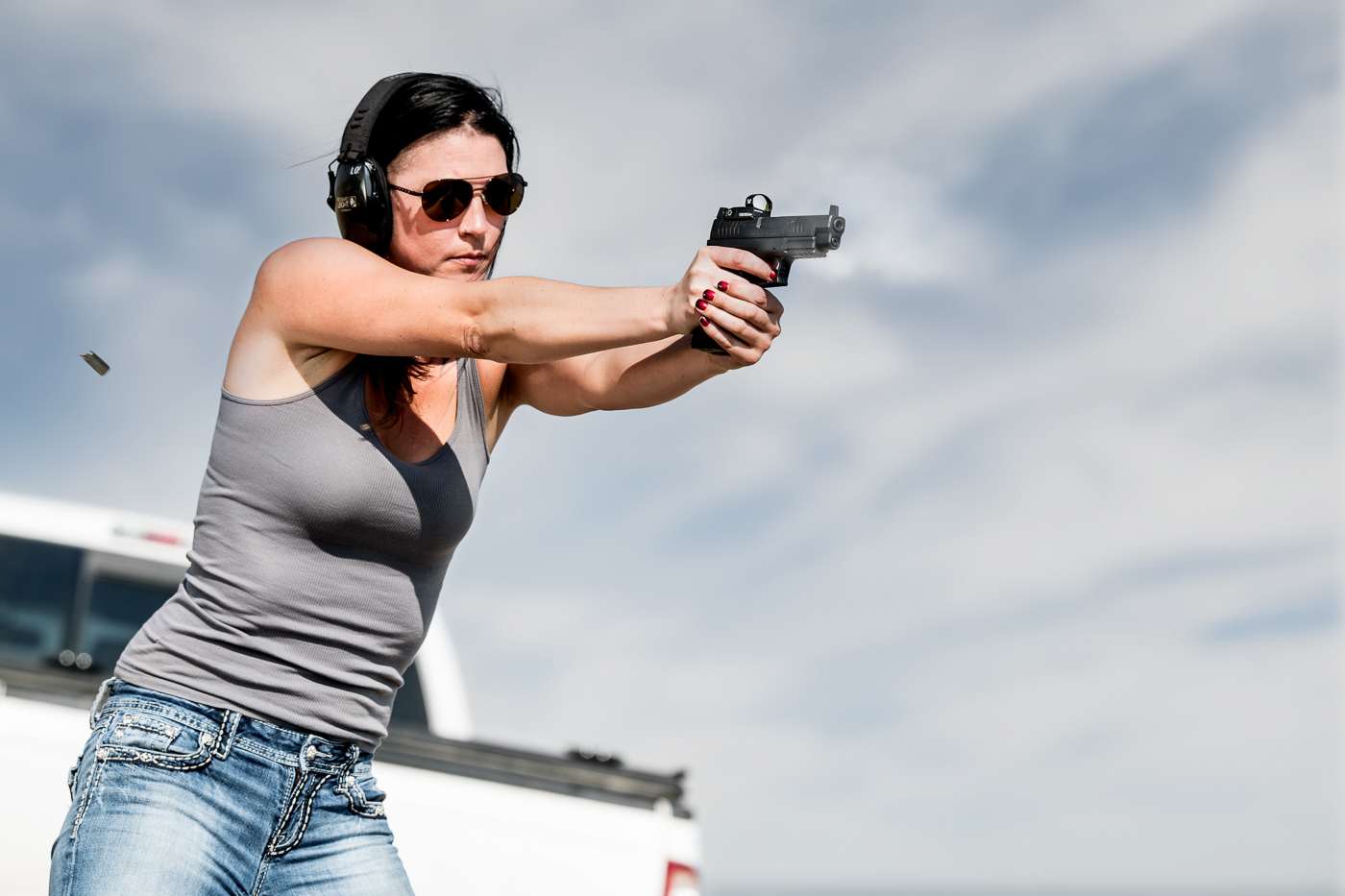 A woman is captured mid-drill as she fires her pistol on an outdoor shooting range. She maintains a firm two-handed grip and proper stance while aiming at her target. The scene shows the active portion of firearms training, where controlled live fire builds accuracy and defensive awareness. Range equipment and target stands are visible in the background, creating a structured training environment. This practice session connects to two self-defense shooting drills that emphasize speed, movement, and using cover. The focus on her technique highlights how shooters gain confidence through repetition and safe habits.