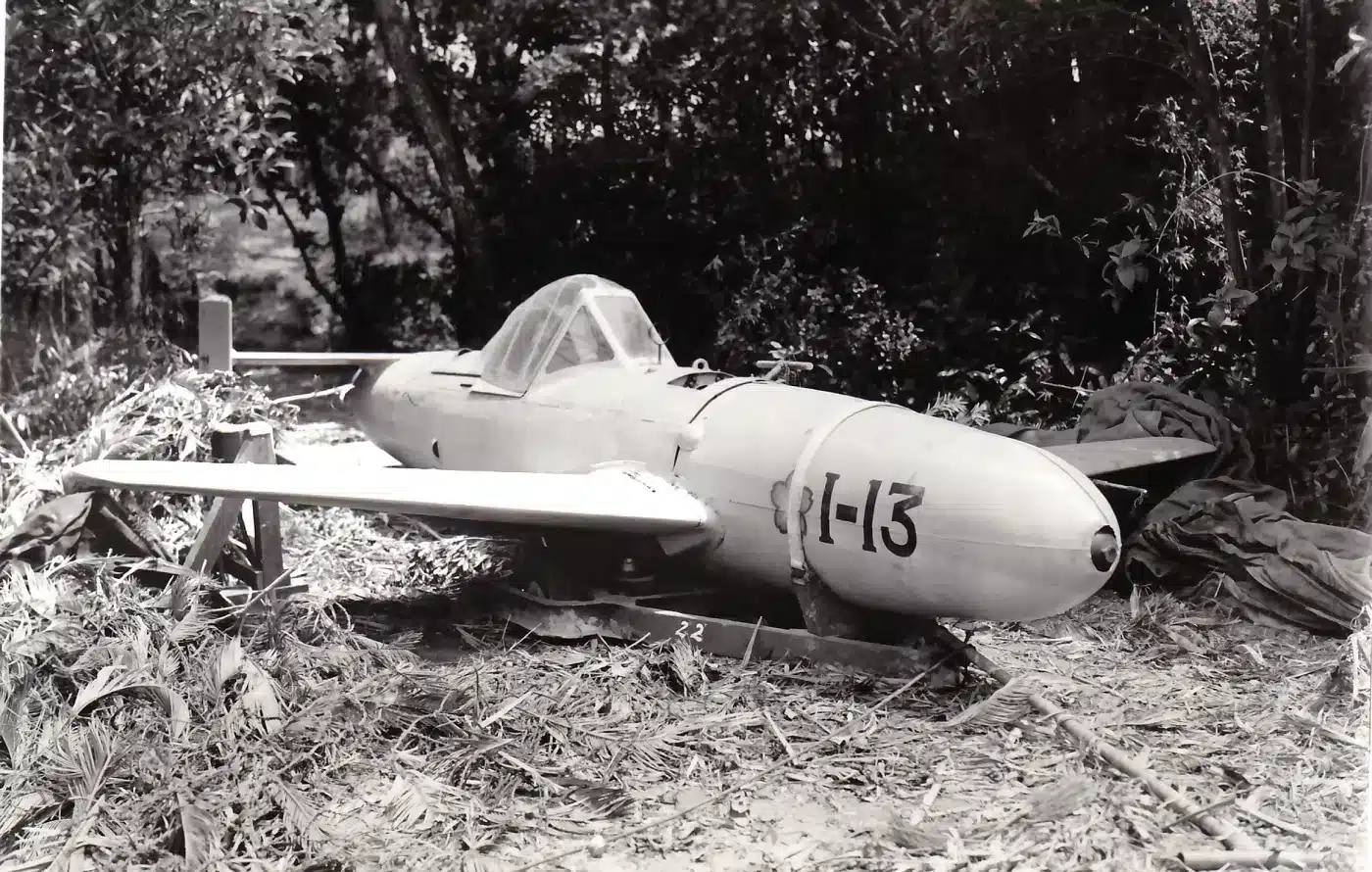 A detailed photograph shows an intact Yokosuka MXY-7 Ohka (“Baka”) rocket-powered kamikaze aircraft captured by U.S. forces on Okinawa in 1945. The small, streamlined fuselage rests on the ground, painted light gray with Japanese markings still visible on the tail and wings. The single-seat cockpit canopy is closed, and the nose section contains the explosive warhead compartment. Several U.S. servicemen stand nearby, examining the captured weapon. The photo highlights the crude yet deadly design of Japan’s manned rocket bombs used in suicide attacks against Allied ships. It’s a rare example of a Baka recovered intact, illustrating the technology behind Japanese Kamikazes in World War II.
