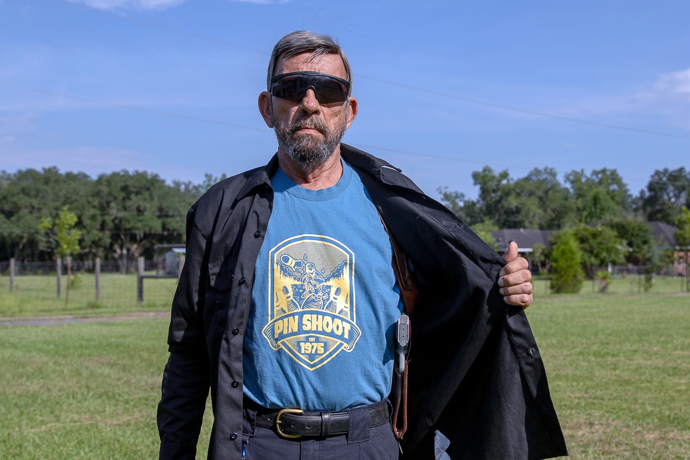 The image shows a man wearing a concealed shoulder holster, often referred to as a Fudd holster, partially covered by a light jacket. The open garment reveals the harness straps and the holstered firearm positioned under the arm. When the jacket is closed, the holster becomes effectively concealed, demonstrating the importance of a proper cover garment. The man’s clothing is casual, showing that this setup can be used in everyday environments. Lighting is neutral, and the background is plain, drawing attention to the holster and clothing interaction. This photo illustrates a key point of the article  —  shoulder carry requires the right outer layer for concealment and discretion. It also reinforces the idea that traditional designs still fit within modern concealed-carry wardrobes.