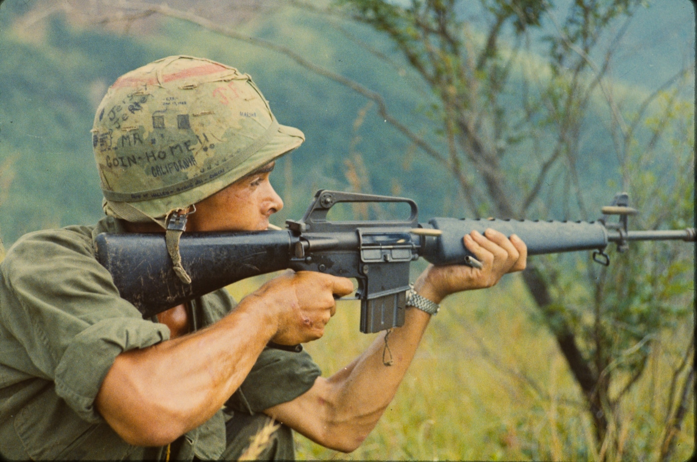 101st Airborne Division soldier fires M16A1 rifle during Vietnam War combat operation in 1967 showing original A1 variant before M16A2 modifications were implemented. The M16A1 rifle features 20-inch barrel with 1:12 twist rate designed for lightweight 52-grain M193 ball ammunition at close engagement distances. Three-prong duck bill flash hider visible on A1 model was prone to catching vegetation and kicking up debris when shooting prone unlike later A2 bird cage design. Triangular handguard configuration on M16A1 used separate left and right components replaced by round interchangeable handguards on A2 variant. Short buttstock length of pull on A1 model was extended in M16A2 design for better shooting ergonomics and shoulder retention. Flip aperture rear sight on M16A1 offered two range settings but lacked adjustability compared to click-adjustable windage and elevation sights on M16A2. Full automatic selector setting on A1 fire control group was changed to three-round burst in M16A2 after Vietnam experience showed full auto wasted ammunition. Soldier demonstrates M16A1 rifle-length gas system and lightweight characteristics that made it maneuverable in jungle warfare versus heavier M14 rifle but revealed need for improvements incorporated into M16A2.
