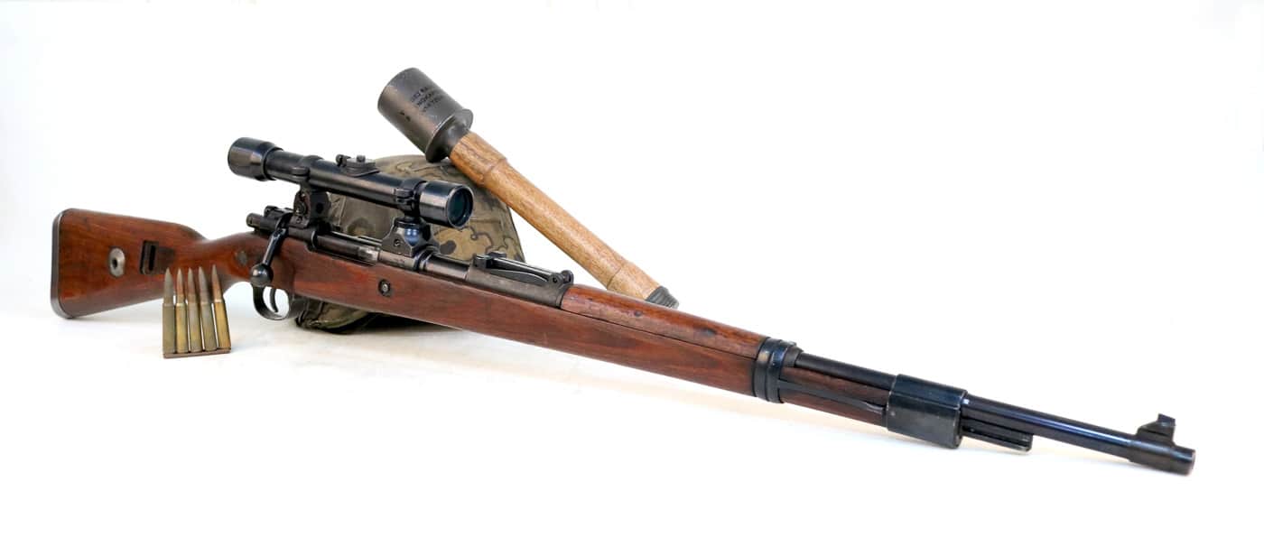 A German Karabiner 98k bolt-action rifle equipped with a ZF41 telescopic sight displayed alongside period-correct Wehrmacht equipment including a steel stahlhelm helmet, stripper clip of 7.92x57mm Mauser ammunition, and Model 24 Stielhandgranate stick grenade. The Kar98k shows the ZF41 scope mounted to the rear sight base, a 1.5X long eye relief optic that approximately 100,000 units were produced during World War 2 though it proved less effective than other German military sights. The stripper clip holds five rounds of 7.92x57mm Mauser s.S. Patrone ammunition used to rapidly charge the rifle's internal magazine from the top of the receiver. German infantry carried multiple stripper clips in ammunition pouches along with grenades, entrenching tools, gas masks, and other combat equipment during operations from 1939 through 1945. The coal scuttle helmet or stahlhelm represents the iconic German military headgear that protected Wehrmacht and Waffen SS soldiers throughout World War 2 on all fronts. The Model 24 stick grenade with its distinctive wooden handle became one of the most recognizable German weapons of the war, used extensively in both offensive and defensive operations. This collection of Mauser rifle accessories and German military equipment shows the typical combat loadout for Wehrmacht infantry armed with the Karabiner 98 kurz standard issue rifle. The ZF41 optical sight mounted on this Kar98k proved less popular than the 4X ZF4 scope due to its small size and limited eye relief that made aimed shooting difficult, though the concept of a low-power combat optic was sound for its era.