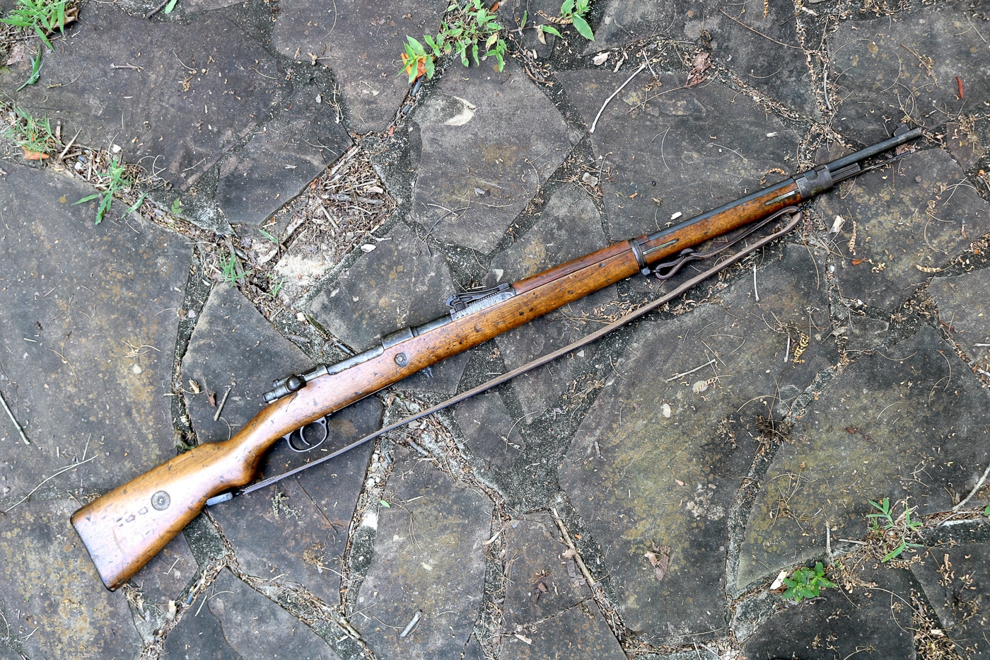 German Gewehr 98 bolt-action rifle displayed with original military leather sling attached showing complete field configuration carried by Imperial German infantry during World War I. The Gew 98 rifle sling connects through metal swivel points mounted on forward barrel band and rear stock allowing soldiers to carry the 9-pound rifle over shoulder during marches and trench movements. This 7.92x57mm Mauser rifle with sling demonstrates standard German military equipment issue where the leather carrying strap proved essential for infantrymen hauling the 49-inch long G98 through battlefield conditions. The sling system on this Gewehr 98 also provided shooting support when soldiers used hasty sling techniques for improved accuracy with the bolt-action rifle. The complete rifle with sling represents typical configuration that millions of German soldiers including runners, infantrymen, and trench fighters carried from 1914 through 1918 during Great War combat operations.