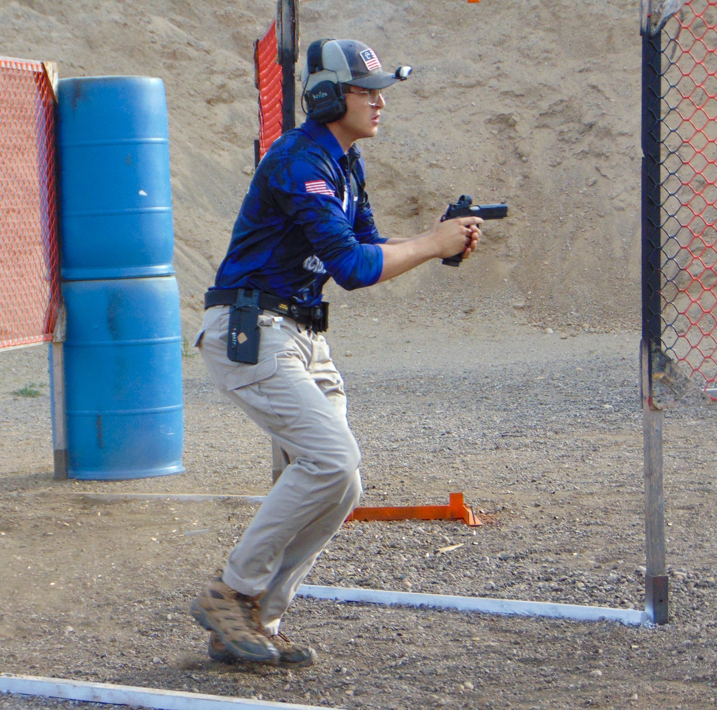 A Hillsdale Chargers shooting team member actively competes during a stage at the 2025 USPSA Collegiate Action Pistol Championships held at their home range facility. The Hillsdale College competitive shooter engages multiple targets with their handgun while moving through the course of fire at the John A. Halter Shooting Center. This Hillsdale action pistol team competitor demonstrates proper gun handling and shooting technique during one of the Springfield Armory sponsored stages. The Hillsdale marksmanship athlete wears eye and ear protection while executing their run through the practical shooting course layout. Hillsdale College pistol team members like this shooter competed in either Limited or Limited Optics divisions using platforms like the 1911 DS Prodigy. The Hillsdale Chargers competitive shooting athlete represents a program that finished second in both team divisions against universities including Texas A&M and Naval Academy. This Hillsdale shooter participates in the type of action pistol stage that requires speed, accuracy, and tactical movement skills valued in USPSA collegiate competition. The Hillsdale team competitor's performance contributes to overall team scores that have established their Michigan college shooting program as a premier destination for university-level practical shooting sports championships.