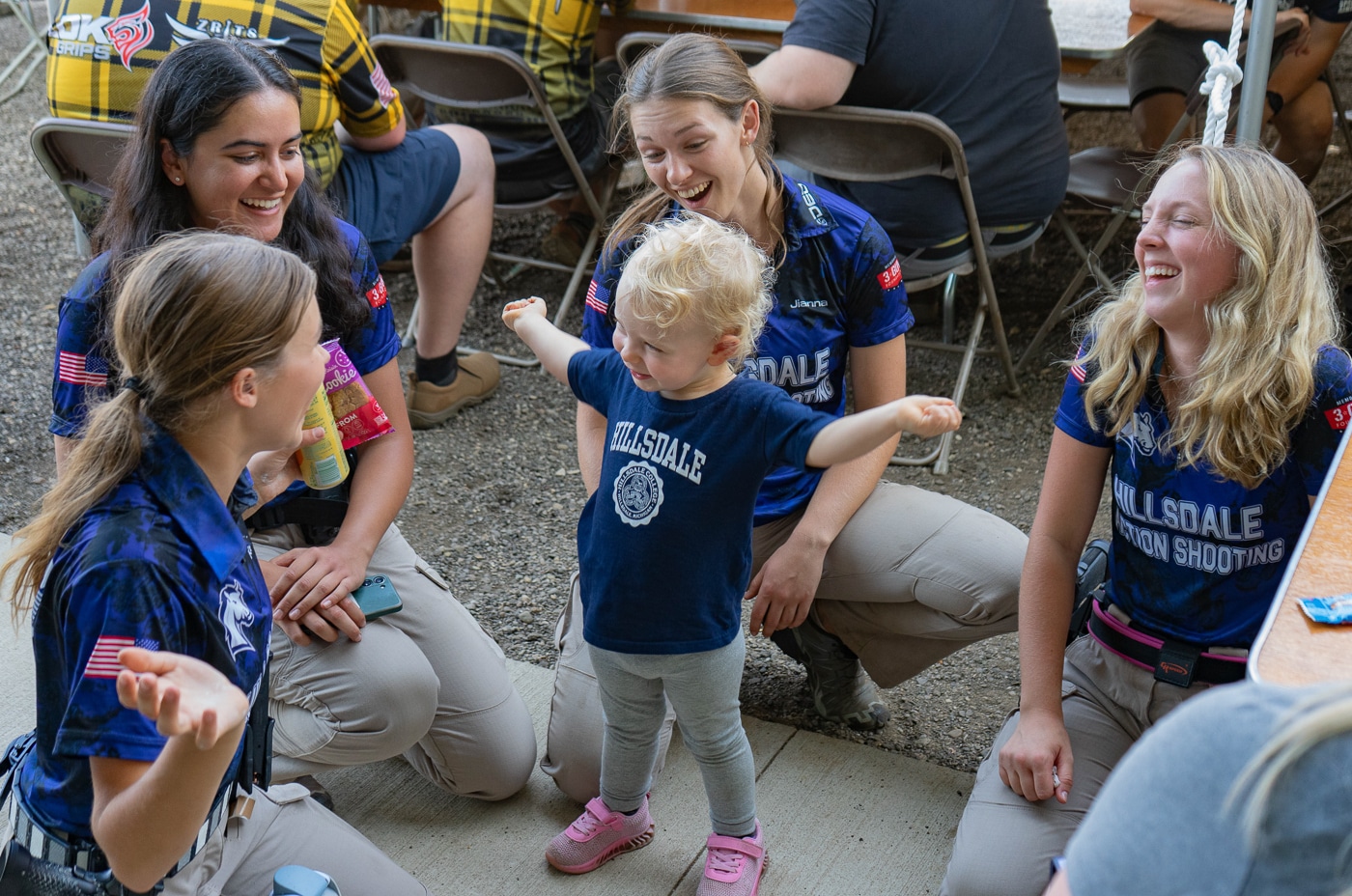 A toddler interacts with members of the Hillsdale Chargers shooting team during breaks at the 2025 USPSA Collegiate Action Pistol Championships held at the John A. Halter Shooting Center. The young child enjoys spending time with Hillsdale College competitive shooting athletes who demonstrate the welcoming and family-friendly nature of collegiate action pistol events. Hillsdale marksmanship team members engage with the toddler while their families and supporters observe the competition at the Roger Ailes Action Pistol Shooting Center in Michigan. The Hillsdale Chargers pistol competitors show that university shooting sports attract multigenerational support including very young fans who attend with parents or supervising adults. Hillsdale College action pistol team athletes take time between stages to interact with young spectators which helps introduce children to competitive shooting sports early. The toddler represents the future of collegiate USPSA competition as Hillsdale shooting program members encourage younger people to learn about marksmanship and practical shooting. Hillsdale Chargers competitive shooters demonstrate the approachable nature of team members who are willing to talk with spectators of all ages at their home facility matches. The young fan's presence at the Hillsdale College shooting team competition shows how the program welcomes families and creates an environment where children can safely observe university-level action pistol sports.
