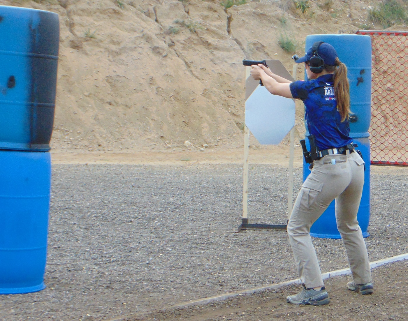 A woman from the Hillsdale Chargers shooting team fires her pistol at targets during a competitive stage run at the 2025 USPSA Collegiate Action Pistol Championships in Michigan. The Hillsdale College competitive shooting athlete moves through the course while engaging cardboard targets set up across the shooting bay at the Roger Ailes Action Pistol Shooting Center. This Hillsdale action pistol team woman demonstrates the shooting fundamentals and stage strategy that helped women competitors dominate the Limited division at their home facility. The Hillsdale marksmanship woman shooter wears safety equipment including eye and ear protection while competing in the timed practical shooting match against teams from Texas A&M, Naval Academy, and Virginia Tech. Hillsdale Chargers pistol team women like this competitor contributed to second place team finishes with standout individual performances including Kayla Mullin's first place Limited HOA score. The Hillsdale College shooting woman engages multiple targets with accuracy and speed that characterizes university-level USPSA action pistol competition where women athletes compete equally alongside male shooters. This Hillsdale competitive shooting athlete represents a program where women shooters Clara Bozzay, Jianna Coppola, Kayla Mullin, and Cristina Lewis have established themselves as top competitors in collegiate marksmanship. The Hillsdale woman competitor's stage run at the John A. Halter Shooting Center demonstrates the skill level that makes their program a premier destination for collegiate action pistol sports.