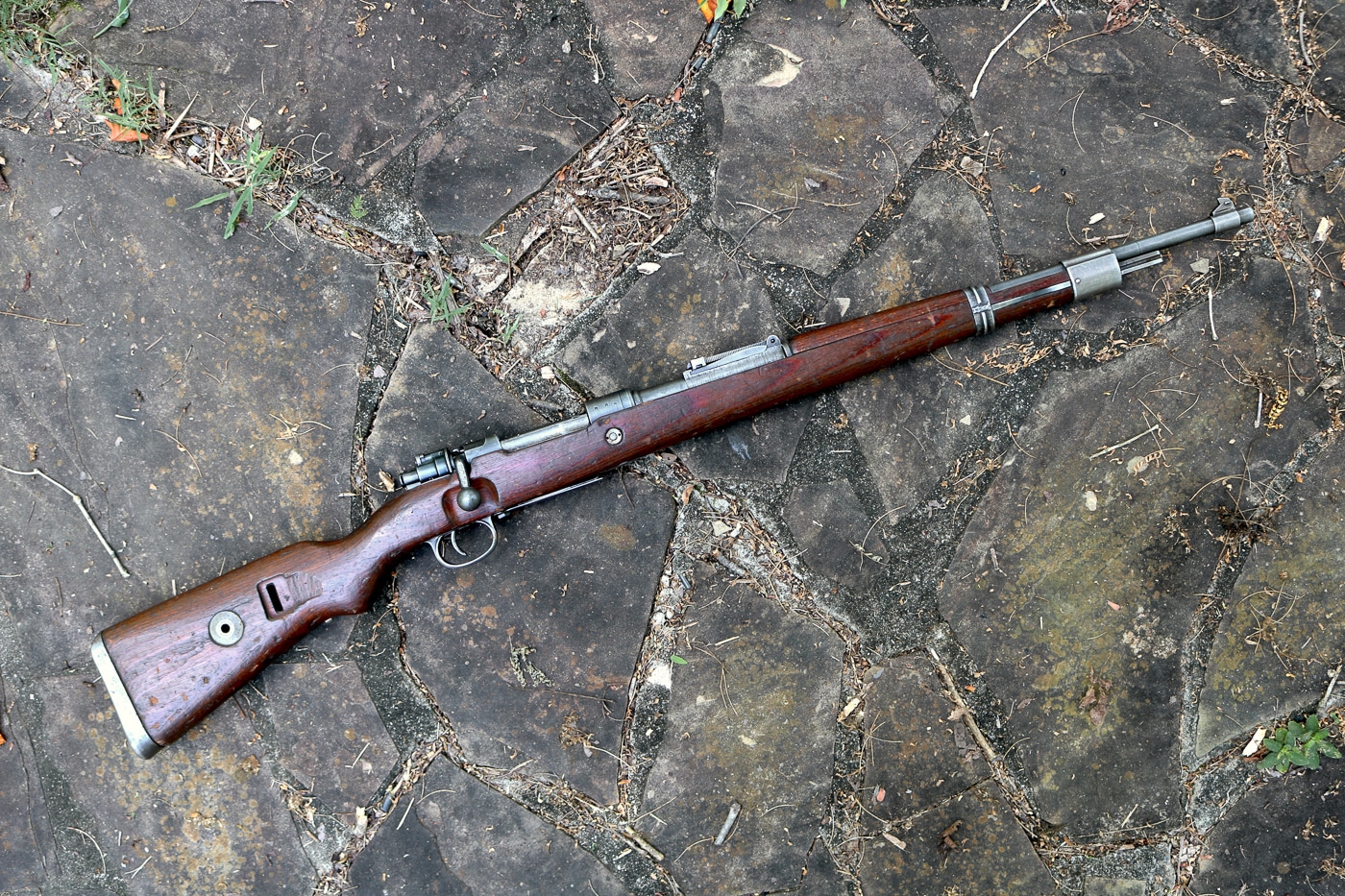 A complete Mauser Karabiner 98k bolt-action infantry rifle representing the primary German service weapon during the Second World War. This Kar98k shows the classic configuration with laminated wood stock, metal receiver, and the shortened 23.6-inch barrel that distinguished the carbine from its Gewehr 98 predecessor. German forces produced approximately 14.6 million of these 7.92x57mm Mauser rifles between 1934 and 1945 for Wehrmacht and SS units fighting across all theaters of World War 2. The rifle features the turned-down bolt handle that allowed mounting of optical sights for sniper use, making the Karabiner 98k a versatile platform for both standard infantry and designated marksmen. The five-round internal magazine loaded via disposable stripper clips provided adequate firepower for bolt-action rifle standards of the era. This German military carbine utilized the controlled-feed M98 Mauser action, one of the strongest and most reliable bolt-action designs ever created for military service. The tangent leaf rear sight and barleycorn front sight combination gave German soldiers accurate aimed fire capability out to effective infantry ranges. Later war production Kar98k rifles like this example featured Parkerized finish instead of traditional bluing and incorporated stamped steel components to speed manufacturing under Allied bombing pressure. The Mauser K98k remains one of the most iconic and recognizable firearms of World War 2, representing German small arms development from the interwar period through the final days of the Third Reich.