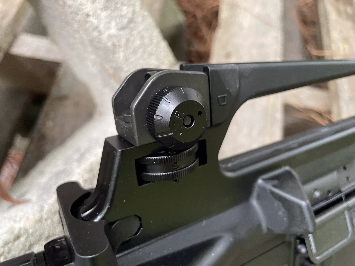M16A2 adjustable rear sight assembly showing major improvement over M16A1 flip aperture design with precision windage and elevation adjustments. Rear sight on A2 features click adjustments ranging from 300 to 800 meters compared to basic two-setting flip aperture on M16A1. Two aperture sizes on M16A2 rear sight include small aperture for daylight precision shooting and large aperture for low-light scenarios. Windage adjustment knob on A2 allows horizontal sight correction lacking on M16A1 flip aperture design. Elevation adjustment drum on M16A2 provides precise range settings from 300 to 800 meters versus simple flip mechanism on A1. Carry handle integrated rear sight on A2 influenced by excellent M14 iron sights requested by Marine Corps after Vietnam War experience. M16A2 sight improvements addressed long-range accuracy needs at 300 to 400 meter modern battlefield distances compared to 25-meter average Vietnam engagements with M16A1. Adjustable rear sight component illustrates key difference between M16A1 and M16A2 variants enhancing downrange performance and precision shooting capabilities similar to M14 characteristics.