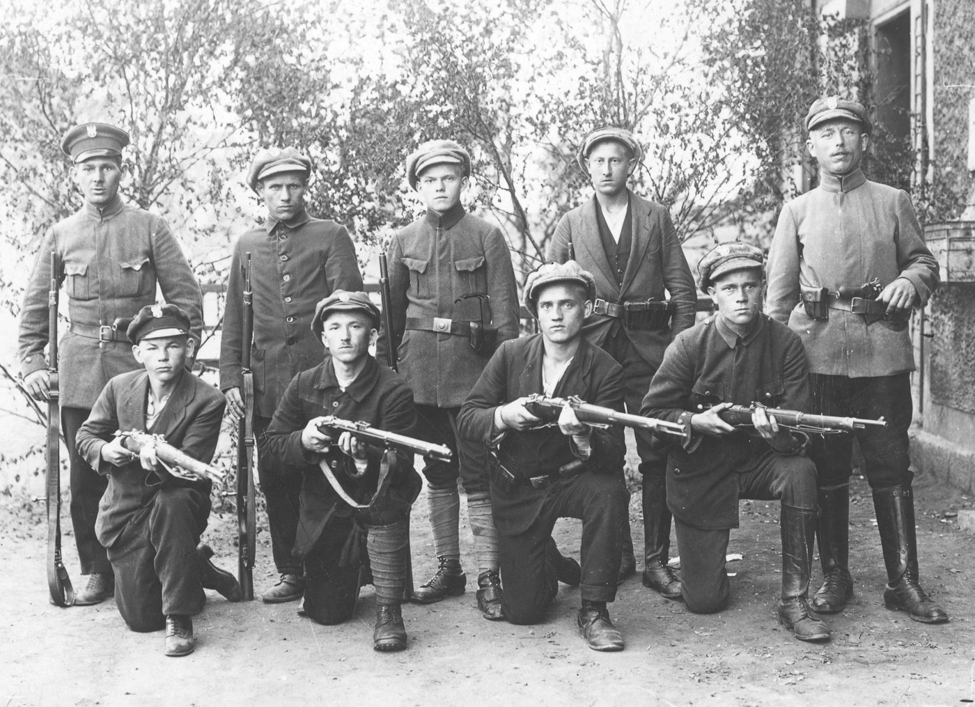 Historical photograph from 1919 showing Polish insurgents armed with German Gewehr 98 bolt-action rifles during combat operations against Bolshevik forces in Eastern Europe. Two kneeling fighters on the right side hold Mauser Gew 98 rifles while the standing insurgent on far right carries a Mauser C96 pistol tucked in his belt. This image demonstrates widespread distribution of German military rifles following World War I when millions of G98 rifles became surplus weapons used by various European forces. The Polish fighters display the 49-inch long Gewehr 98 rifles with their distinctive straight bolt handles and 29-inch barrels. These 7.92x57mm Mauser rifles served Polish irregular forces during the Polish-Soviet War when captured German arms became essential weapons for Eastern European militaries and resistance groups fighting against communist expansion.