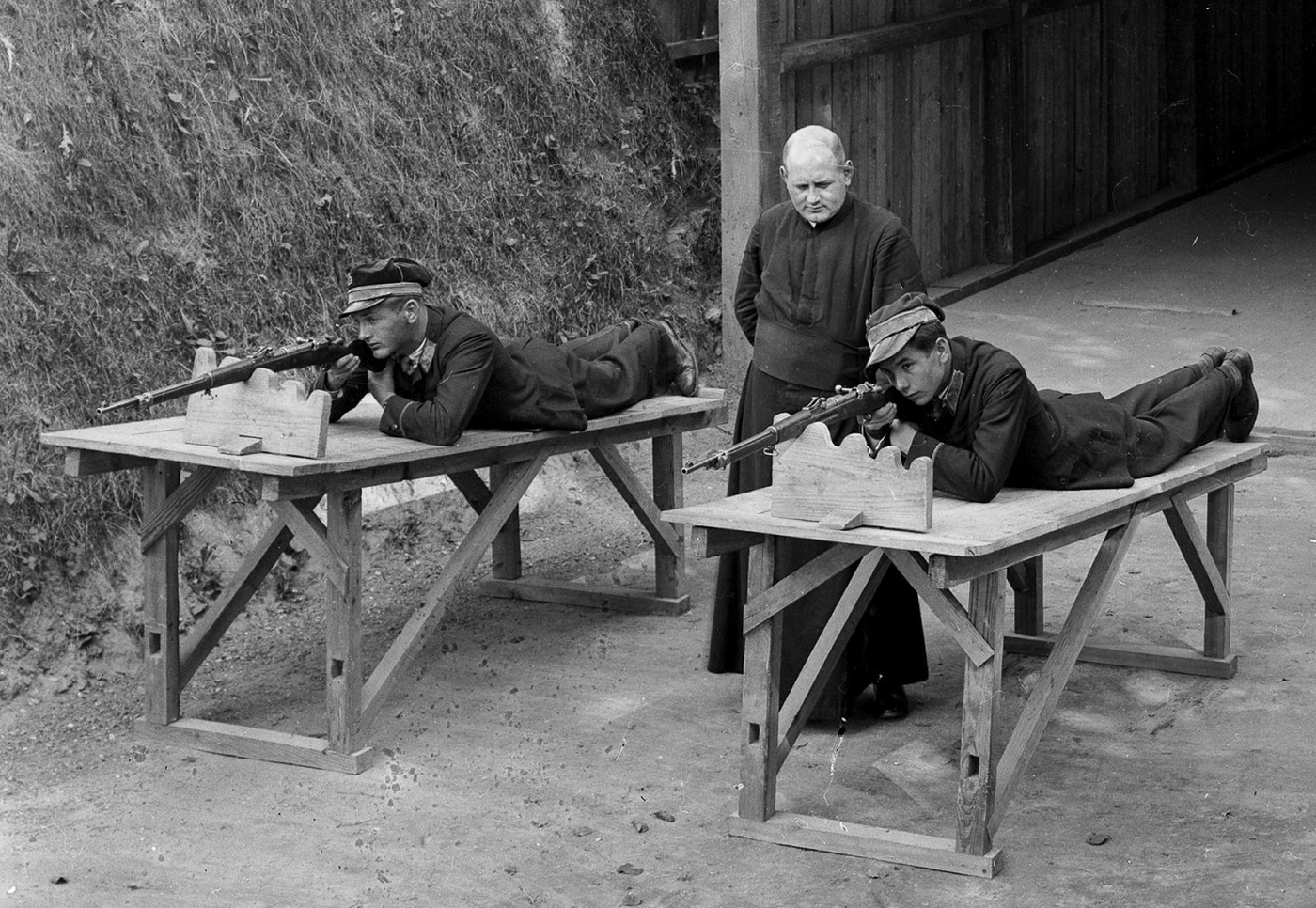 Historical photograph showing Roman Catholic priest providing marksmanship instruction to young Polish men using German Gewehr 98 bolt-action rifles demonstrating civilian firearms training programs in Poland during interwar period following World War I. The priest instructs youth in proper handling of Gew 98 rifles or Polish Mauser wz. 98 variants chambered in 7.92x57mm Mauser that became widely available throughout Eastern Europe after millions of German military rifles flooded region as war surplus. These young Polish men learning to shoot with Gewehr 98 rifles represent community preparation for military service when Poland faced threats from both Germany and Soviet Union during 1920s and 1930s leading to World War II. The marksmanship training using reliable controlled-feed Mauser action G98 rifles shows how Catholic Church participated in preparing Polish youth for national defense using same bolt-action rifle design that equipped German forces during Great War and would later arm Polish military units.