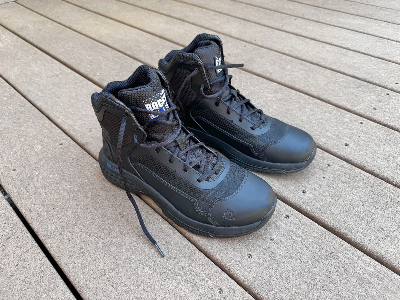 Rocky Code Blue Public Service boots shown in outdoor setting illustrating tactical footwear designed for law enforcement patrol officers with six-inch height and breathable construction. Police duty boots feature mixed-material upper combining cushiony mesh panels with sewn and cemented TPU thermoplastic polyurethane overlays creating durable yet comfortable patrol boot. Non-waterproof tactical boots include EVA ethylene-vinyl acetate Rebound midsole providing shock absorption and elastomeric cushioning for extended wear during patrol shifts. Oil-resistant rubber outsole displays lateral grooves and concentric tread patterns preventing hydroplaning on wet pavement and providing grip on varied terrain from urban streets to drainage ditches. Rocky Boots affordable law enforcement footwear offers professional appearance with ability to accept Kiwi shoe polish for high-gloss shine required for Class A uniform inspections. Public safety tactical boots tested by experienced K9 officer with 25 years patrol experience and 12,000 miles on-duty walking running and climbing. Budget tactical boot option under $100 provides patrol officers security professionals and first responders with presentable durable footwear handling city hall meetings field searches and emergency response scenarios. Six-inch non-metallic boots include oval laces padded collar tongue and fiberglass shank construction eliminating traditional leather jump boot Wellington boot designs for modern lightweight patrol footwear.