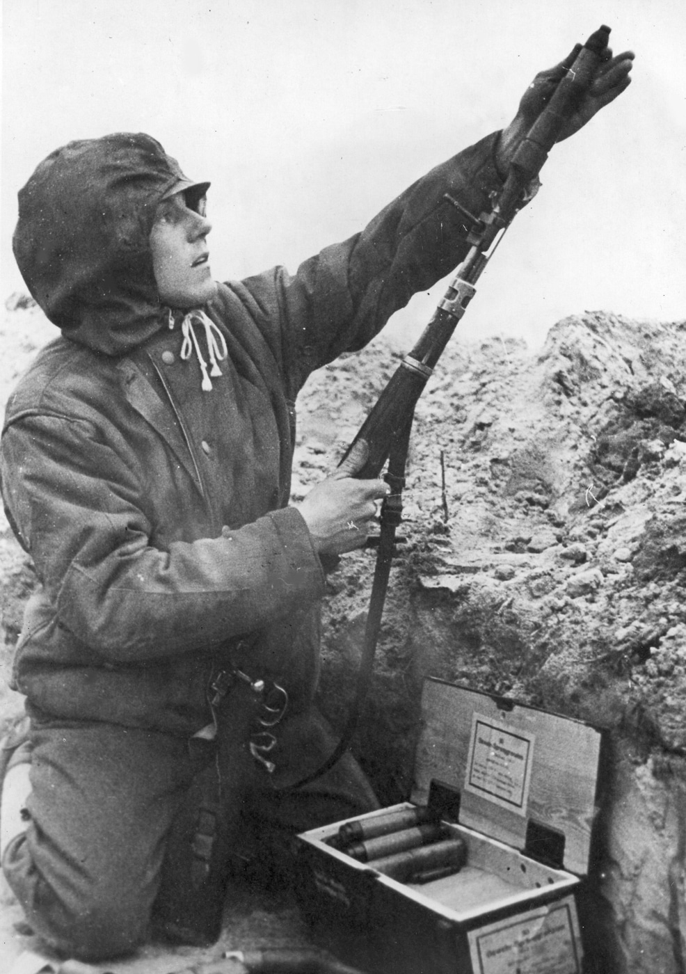A German Wehrmacht soldier in combat uniform loads a Gewehr-Panzergranate anti-tank rifle grenade into the Schiessbecher cup-style launcher mounted on his Karabiner 98k bolt-action rifle while fighting from a trench position on the Eastern Front during World War 2. The Kar98k equipped with the Gewehrgranatengerät launcher system gave German infantry the ability to engage armored vehicles, fortifications, and personnel targets at ranges beyond standard rifle fire using specialized grenade projectiles. The soldier demonstrates proper handling technique for rifle grenade operations, inserting the grenade's tail boom into the cup launcher affixed to the muzzle of the 7.92x57mm Mauser rifle. Special grenade launching cartridges with wooden bullets provided propulsion for rifle grenades while automatically priming the explosive warhead upon firing from the Karabiner 98 kurz. Eastern Front combat conditions from 1941 through 1945 saw extensive use of rifle grenades by Wehrmacht troops fighting Soviet forces in trench warfare, urban combat, and defensive positions across Russia and occupied territories. The Schiessbecher launcher cup attached to the Kar98k muzzle using a clamp system that could be removed when standard rifle fire was required, giving German infantrymen flexibility in combat roles. German soldiers carried multiple rifle grenade types including anti-personnel fragmentation rounds and anti-tank Panzergranate projectiles with shaped charge warheads capable of penetrating light armor. The Gewehrgranatengerät system later adapted to other German weapons including the semi-automatic G98/40, the revolutionary MP44 assault rifle, and the selective-fire FG42 paratroop rifle for broader tactical employment.