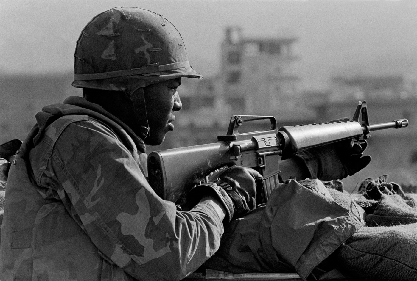 United States Marine Corps guard armed with M16A1 rifle at Beirut International Airport bunker position shows A1 variant before M16A2 replacement in 1983. The M16A1 rifle features three-prong duck bill flash hider design that collected debris unlike enclosed bird cage flash suppressor on M16A2 variant. Triangular handguard system on A1 model used weaker material than reinforced polymer round handguards on M16A2 design. Short buttstock with smooth plate on M16A1 lacked extended length of pull and textured surface of M16A2 stock for better shoulder retention. Marine demonstrates M16A1 flip aperture rear sight with basic range settings compared to fully adjustable windage and elevation sights on A2 from 300 to 800 meters. Full-automatic fire selector on A1 fire control group visible differs from three-round burst capability on M16A2 requested by Marines. Barrel twist rate of 1:12 inches on M16A1 optimized for 52-grain M193 ammunition inadequate for heavier 62-grain M855 rounds requiring 1:7 twist on M16A2. Peacekeeping mission illustrates M16A1 service during transition period when Marine Corps requested modifications resulting in M16A2 variant with improved long-range accuracy and durability for 300 to 400 meter engagements.