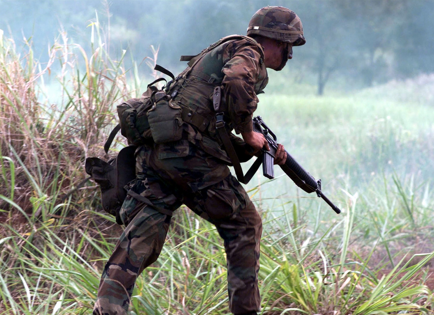 United States Marine Corps infantryman carries M16A2 rifle during combat assault showing differences between M16A1 and M16A2 variants in tactical operation. The M16A2 rifle features improved barrel with 1:7 twist rate versus M16A1 1:12 twist for stabilizing heavier M855 ammunition at extended ranges. Marine demonstrates A2 model upgrades including adjustable rear sight with windage and elevation adjustments from 300 to 800 meters compared to basic flip aperture sight on A1 variant. The rifle's bird cage flash hider visible on A2 replaced three-prong duck bill flash hider from M16A1 design. A2 handguard made from stronger polymer material versus triangular A1 handguard shows enhanced durability improvements. Longer stock length of pull and brass deflector for left-handed shooters distinguish A2 from earlier A1 configuration. Three-round burst selector replaced full auto capability from M16A1 for better ammunition conservation. Combat scenario illustrates practical battlefield application of M16A2 modifications requested by Marines based on Vietnam War experience with M16A1 performance issues.