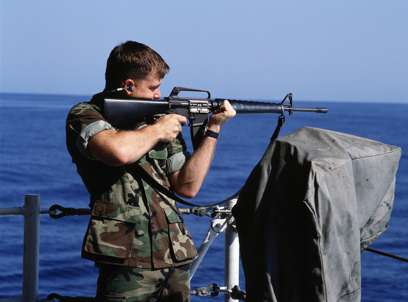 United States Marine fires M16A1 rifle from fantail of USS Kitty Hawk aircraft carrier CV-63 showing A1 variant before M16A2 replacement in 1980s. M16A1 features three-prong duck bill flash hider that collected debris unlike enclosed bird cage flash suppressor on M16A2 variant. Triangular handguard system on A1 made from weaker material than reinforced polymer round handguards on M16A2 design. Short buttstock with smooth plate on M16A1 lacked extended length of pull and textured surface of M16A2 stock for better shoulder retention. Marine demonstrates M16A1 flip aperture rear sight with basic range settings compared to fully adjustable windage and elevation sights on A2 from 300 to 800 meters. Full-automatic fire selector on A1 visible differs from three-round burst capability on M16A2 requested by Marines for better accuracy. Barrel twist rate of 1:12 inches on M16A1 optimized for 52-grain M193 ammunition inadequate for heavier 62-grain M855 rounds requiring 1:7 twist on M16A2. Shipboard scenario illustrates M16A1 lightweight characteristics and rifle-length gas system with minimal recoil retained in M16A2 while incorporating enhanced sights, stronger materials, and improved long-range performance at 300 to 400 meter distances.