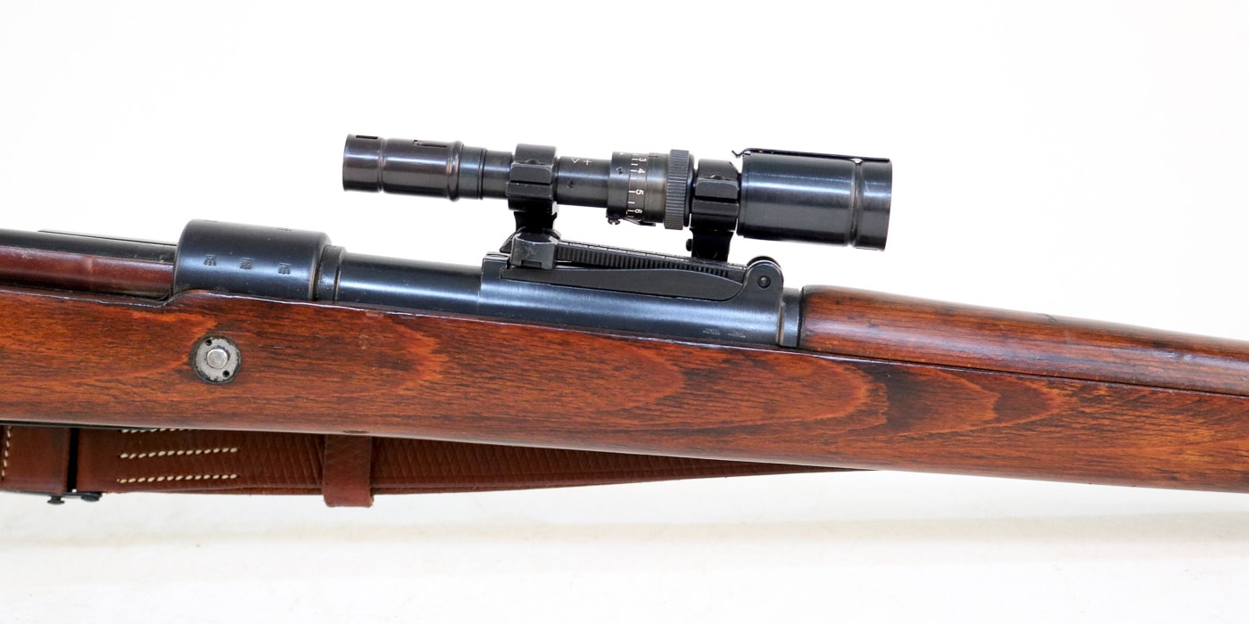 Close-up view of a ZF41 telescopic sight mounted on a Karabiner 98k Mauser rifle showing the distinctive 1.5X long eye relief optic that attached to the rear sight base of the German bolt-action infantry weapon during World War 2. The ZF41 scope represents one of three major optical sights used on the Kar98k, with approximately 100,000 units produced between 1941 and 1945 for Wehrmacht and Waffen SS use across all combat theaters. This compact optic mounted directly to the existing rear sight base of the 7.92x57mm Mauser rifle without requiring special machining or gunsmithing, making it economical to produce and install compared to the ZF4 and ZF39 scopes that needed dedicated mounting rails. The small objective lens and tube diameter of the ZF41 created a restricted sight picture that German soldiers compared to looking through a narrow tube, limiting its effectiveness despite the theoretical advantage of 1.5X magnification. German military doctrine attempted to provide standard infantry with modest optical enhancement through the ZF41 system, though the limited eye relief and small field of view made rapid target acquisition difficult during combat operations. The ZF41 telescopic sight on the Karabiner 98 kurz proved less popular than iron sights or the superior 4X ZF4 scope that designated marksmen preferred for actual sniper and precision shooting applications. This long eye relief optic allowed German riflemen to maintain normal shooting position and cheek weld on the Kar98k stock while using the scope, unlike conventional scopes that required closer eye positioning to the ocular lens. The ZF41 mounting system's simplicity meant any standard Karabiner 98k could be fitted with the optic in the field without specialized tools or modifications to the receiver, though most soldiers preferred removing the scope and using traditional iron sights for general infantry combat.