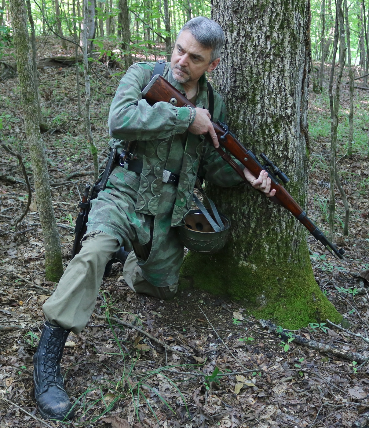 Military firearms author and historian Will Dabbs holds a surplus Karabiner 98k Mauser rifle while wearing period-correct German uniform reproductions that replicate Wehrmacht infantry combat dress from World War 2. The Kar98k bolt-action rifle represents one of millions produced between 1934 and 1945 that served German forces on all fronts throughout the war. Dabbs demonstrates proper handling of the 7.92x57mm Mauser rifle, showing how German soldiers would have carried and presented the Karabiner 98 kurz during infantry operations. The period uniform includes typical Wehrmacht field gray tunic, trousers, boots, and equipment that German riflemen wore while armed with the standard issue Kar98k service rifle. Surplus Mauser rifles remain available to collectors and shooters today through various importers who brought demilitarized and shooter-grade examples from European stockpiles after the war ended. The author's recreation helps modern audiences understand the weight, balance, and ergonomics of the German military rifle that proved both reliable and accurate despite being obsolete compared to semi-automatic designs like the American M1 Garand. Original Kar98k rifles in collector condition command significant prices due to historical interest in World War 2 German firearms, with matching numbers examples and rare variants being especially valuable. The Mauser 98k design remains mechanically sound today, with the controlled-feed bolt action proving so successful that commercial sporting rifles still use variations of the same system developed over a century ago.
