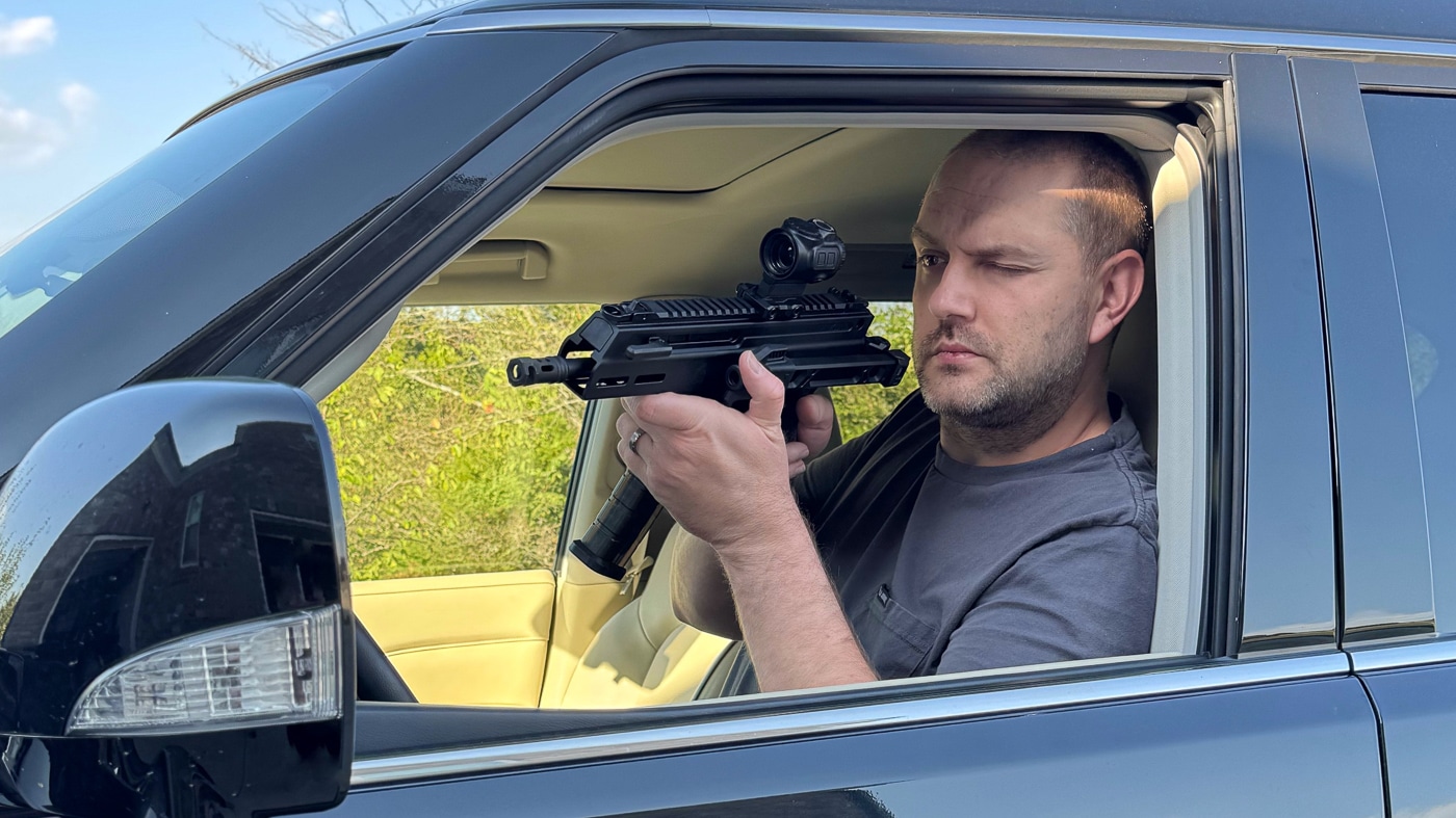 Author demonstrating Springfield Armory Kuna truck gun deployment from vehicle interior showing realistic defensive shooting position and confined space handling. Shooter practicing with Kuna defensive pistol from driver seat illustrating compact design advantages for automotive self-defense scenarios. Springfield Armory truck gun training from vehicle showing roller-delayed pistol performance in tight quarters typical of defensive situations. Kuna 9mm pistol being fired from inside vehicle demonstrating practical truck gun usage and realistic personal protection training. Vehicle interior shooting practice with Springfield Armory Kuna featuring folding brace utility for confined automotive environments. Defensive firearm training showing Kuna truck gun deployment speed and handling from realistic vehicle position during emergency scenarios. Springfield Armory Kuna vehicle defense practice illustrating compact barrel length benefits and maneuverability in tight interior spaces. Author conducting hands-on truck gun training with Kuna pistol from driver position emphasizing real-world defensive preparedness.