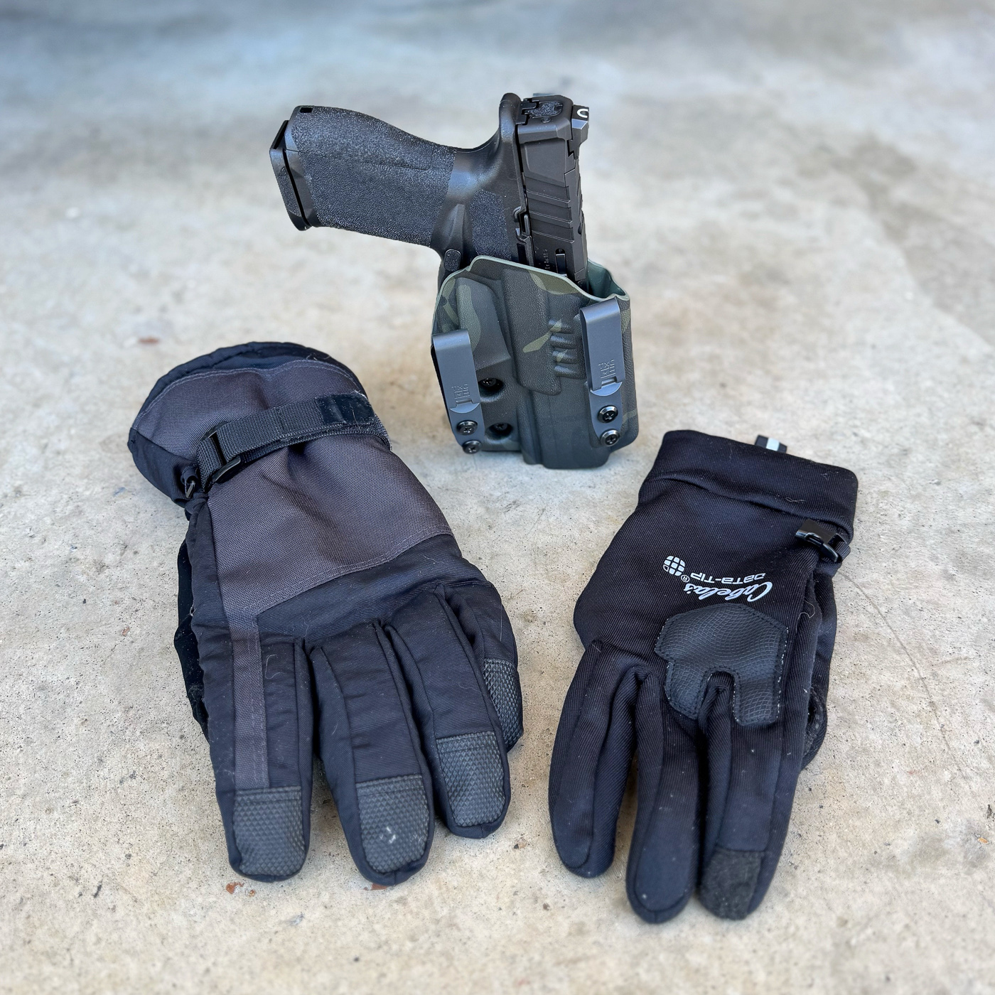 Cold weather shooting gloves designed specifically for concealed carry and firearms handling showing reinforced palms and articulated trigger finger for maintaining trigger guard access and tactile sensitivity in sub-zero temperatures. Photo displays tactical shooting gloves similar to Mechanix Wear or 5.11 Tactical models featuring thin profile that permits proper grip purchase and master grip acquisition on pistol while providing insulation against cold-induced stiffness that impairs fine motor skills. Image shows winter EDC gloves with touchscreen compatible fingertips allowing emergency 911 calls without removal and demonstrating balance between thermal protection and trigger finger dexterity required for defensive scenarios in cold climate conditions. Shooting-specific gloves for concealed carry illustrating design features like flexible knuckles, secure wrist closures, and low-profile construction that accommodates draw stroke and reholstering unlike bulky ski gloves or heavy work gloves that prevent trigger discipline. Cold weather firearm gloves showing merino wool liner alternative or synthetic insulation options that maintain combat grip capability while protecting against hypothermia during extended outdoor carry in northern states or snow belt regions. Tactical gloves for winter concealed carry demonstrating proper equipment selection that supports muscle memory from dry fire practice and draw drills rather than compromising defensive readiness with excessive bulk that blocks trigger guard or prevents safe reholstering. Concealed carry shooting gloves positioned to show palm reinforcement and finger articulation necessary for maintaining muzzle discipline and proper presentation technique when drawing from Kydex holsters or hybrid holsters beneath heavy winter jackets in freezing conditions.