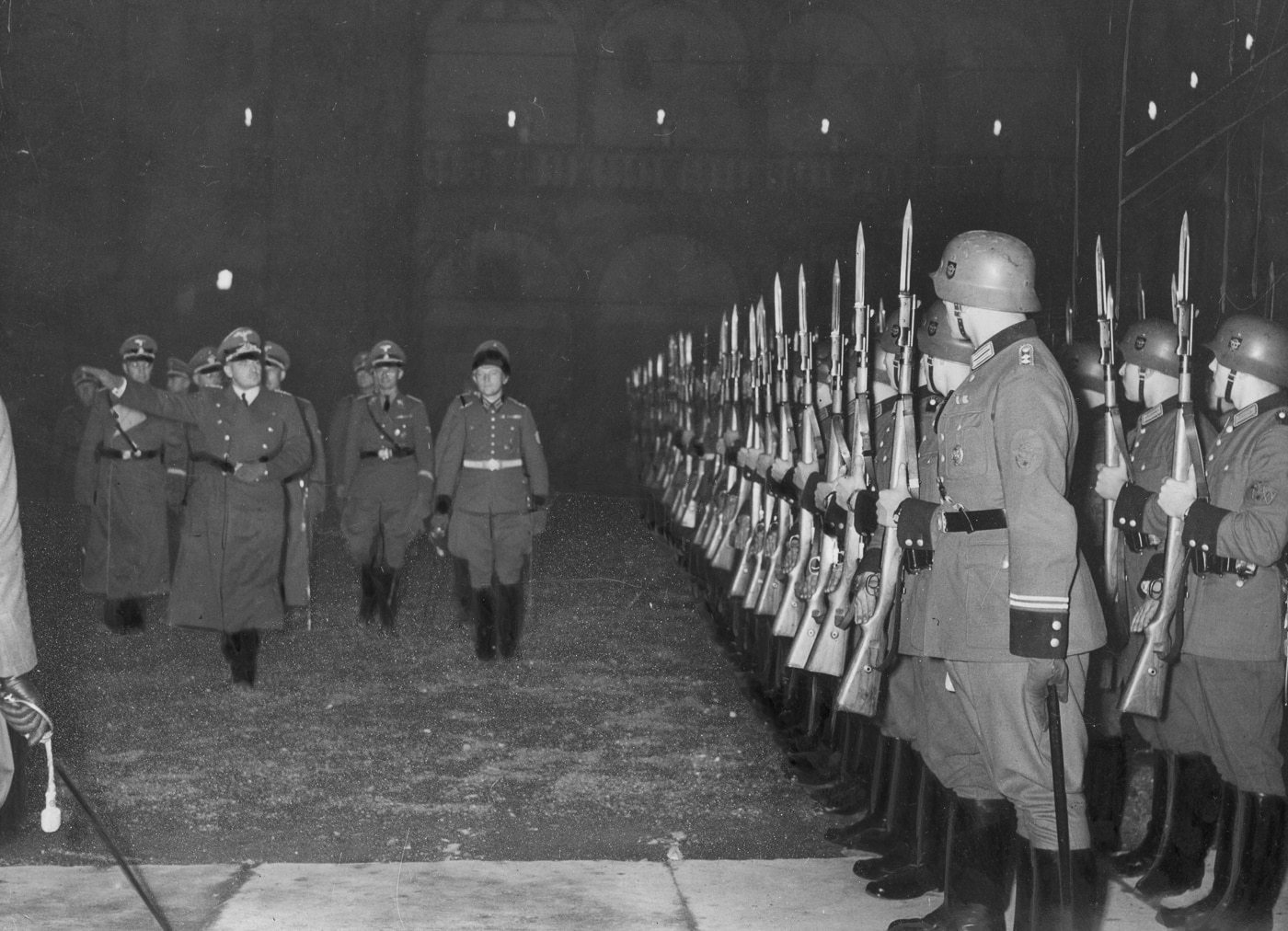 German honor guard troops stand at attention holding Karabiner 98k Mauser rifles during the ceremony where Hans Frank assumed command of the General Government for the Occupied Polish Region following Nazi Germany's conquest of Poland in 1939 during World War 2. The Wehrmacht soldiers carry the standard Kar98k bolt-action rifles in ceremonial presentation position, demonstrating how the 7.92x57mm Mauser service weapon served both combat and formal military functions throughout the Third Reich. Hans Frank served as Governor-General of occupied Poland from 1939 until 1945, overseeing brutal occupation policies that resulted in millions of deaths and earning him a death sentence at the Nuremberg Trials after Germany's defeat. The honor guard's Karabiner 98 kurz rifles represent the standard German infantry weapon that was used not only on battlefields across Europe, Africa, and the Eastern Front but also for ceremonies, parades, and official state functions in occupied territories. German military tradition emphasized formal ceremonies and guard duties, with soldiers carrying their Mauser 98k rifles in pristine condition for official events while the same weapon design saw hard use in combat from Poland in 1939 through Germany's surrender in 1945. The General Government established by Nazi Germany in occupied Poland became the site of horrific war crimes and genocide, with Frank's administration implementing policies that led to the extermination of Polish Jews and the brutal subjugation of the Polish population. Wehrmacht and Waffen SS troops used the Kar98k for honor guard duties across all occupied territories including France, Norway, the Low Countries, the Balkans, and conquered Soviet territories where German military governors held power. The ceremonial use of the Karabiner 98k bolt-action rifle contrasts sharply with its primary role as the standard combat weapon that millions of German soldiers carried into battle throughout six years of total war that devastated Europe and resulted in tens of millions of deaths.