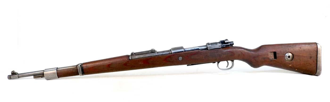Left side view of a German Karabiner 98k Mauser bolt-action rifle showing the turned-down bolt handle, receiver markings, cleaning rod, and overall profile of the standard Wehrmacht infantry weapon that served throughout World War 2 from 1934 to 1945. The Kar98k left receiver wall displays manufacturer codes, production dates, and inspection stamps that German arsenals applied during manufacturing and acceptance testing of the 7.92x57mm service rifles produced by multiple contractors across Nazi Germany. The turned-down bolt handle distinguishes the Karabiner 98 kurz from the earlier Gewehr 98 design, reducing cycling effort and providing clearance for mounting telescopic sights on sniper variants equipped with ZF4, ZF39, or ZF41 optics. A bolt release latch visible on the left side of the receiver allows German soldiers to disassemble the rifle for cleaning and maintenance by retracting the bolt, pulling up on the latch, and removing the bolt assembly to the rear without tools. The short threaded cleaning rod secured underneath the barrel via a retaining band provided Wehrmacht infantry with basic field maintenance capability, allowing soldiers to clean the bore without carrying separate cleaning equipment in their combat loadouts. The Mauser 98k left side profile shows the laminated plywood stock that became standard after 1937, replacing earlier one-piece wooden stocks with more robust construction that resisted cracking and weather damage better than solid wood furniture. The receiver ring and magazine well are visible from this angle, showing the controlled-feed design of the Mauser M98 action where the extractor snaps over the cartridge rim as rounds feed from the five-round internal magazine. German manufacturers produced approximately 14.6 million Karabiner 98k rifles between 1934 and 1945 at various arsenals and contracted facilities, with each weapon bearing specific marking codes that identify the manufacturer, production year, and acceptance proofs applied during quality control inspection.