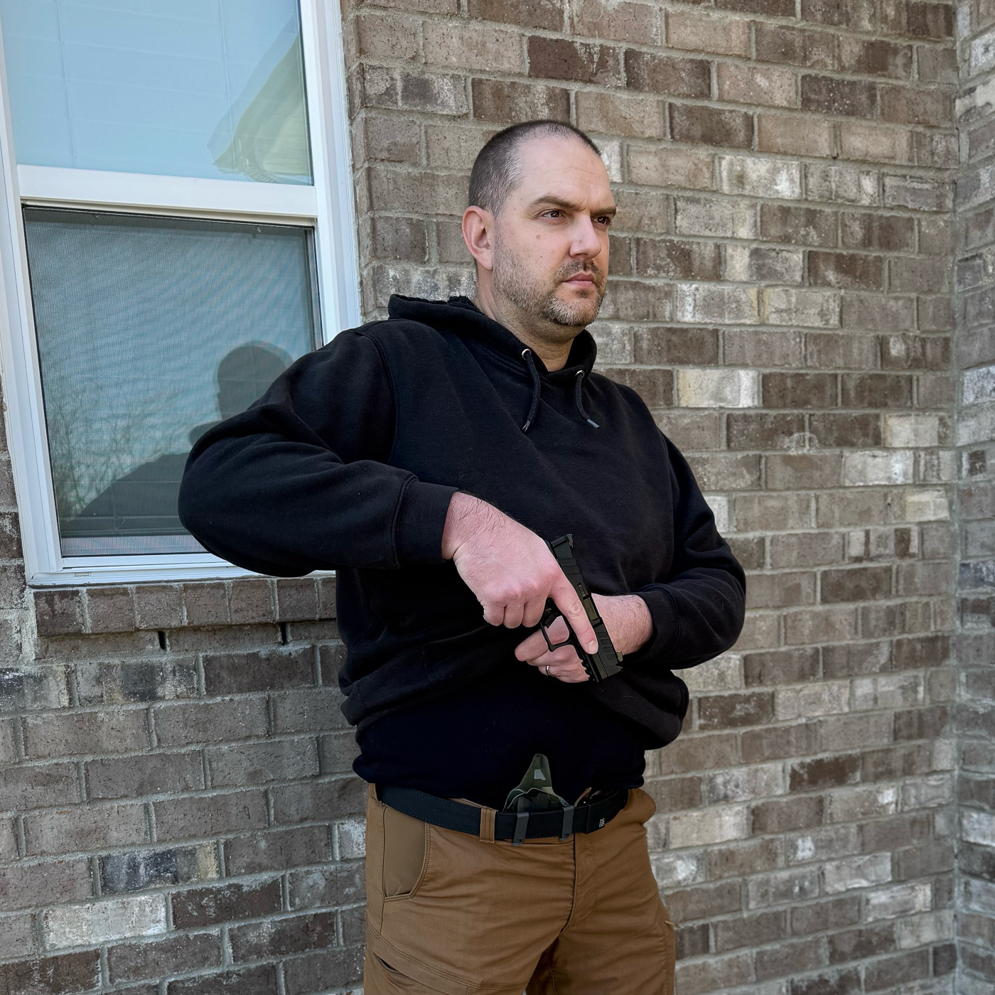 Concealed carrier demonstrating appendix carry draw technique by lifting hoodie sweatshirt to access pistol in AIWB holster positioned at front centerline showing popular winter EDC setup for younger carriers and casual clothing environments. Photo illustrates proper garment sweep modification for pullover-style cover garments as shooter pulls hoodie upward with support hand while establishing master grip on pistol in appendix position holster rather than traditional strong side carry location. Man executing AIWB draw from concealed Kydex holster beneath hoodie sweatshirt revealing how inside the waistband appendix carry requires different clearing motion than jacket-based concealment for cold weather concealed carry situations. Image shows tactical draw from appendix holster under pullover hoodie demonstrating accessibility challenges that sweatshirts and pullovers introduce compared to zip-up jackets or button-front cover garments for defensive readiness scenarios. Concealed carry presentation from AIWB position beneath hoodie illustrates why appendix carry practitioners must practice draw drills with various cover garment types including sweatshirts that cannot be swept aside like traditional flannel shirts or CCW jackets. Shooter drawing handgun from appendix inside the waistband holster after lifting front hem of hoodie sweatshirt showing grip purchase technique and trigger guard access when carrying concealed in casual winter clothing rather than tactical apparel from 5.11 or Vertx brands. Appendix carry draw stroke from beneath hoodie demonstrates gun belt placement and holster retention considerations for AIWB setup where lifting fabric upward rather than sweeping sideways affects draw time and muscle memory requirements for winter concealed carry training.