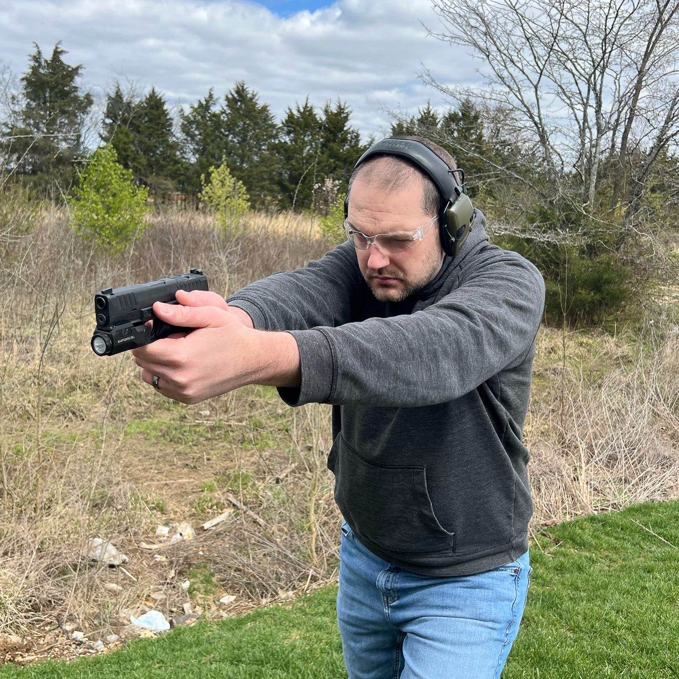 Concealed carrier conducting dry fire practice and draw drills wearing sweatshirt or hoodie with holstered pistol demonstrating cold weather concealed carry training essential for maintaining defensive readiness and muscle memory with winter clothing layers. Photo shows shooter training with actual EDC setup including cover garment and holster configuration practicing presentation technique and garment sweep fundamentals required when carrying concealed firearms beneath pullover sweatshirts or hoodies during cool weather conditions. Man executing draw stroke practice in sweatshirt illustrating importance of integrating winter clothing into regular training routine whether practicing with Glock, Springfield Armory, Sig Sauer P365, or Smith & Wesson M&P pistols in Kydex holsters or hybrid holsters at appendix carry or strong side positions. Cold weather firearms training session showing concealed carry practitioner rehearsing grip purchase, trigger guard access, and reholstering techniques while wearing actual winter EDC clothing rather than training only in summer attire that doesn't replicate fabric interference challenges. Image demonstrates tactical training methodology for winter concealed carry as shooter practices draw drills with sweatshirt cover garment to identify printing issues, accessibility problems, and draw time increases that bulkier clothing introduces compared to t-shirt carry during warmer months. Concealed carry training photo showing defensive readiness preparation through repeated practice of garment clearing motions, combat grip establishment, and presentation fundamentals while wearing winter layers that affect fine motor skills and range of motion during draw stroke execution. Shooter conducting live fire training or dry fire practice in cold weather clothing setup including sweatshirt or fleece jacket demonstrating why USCCA, NRA instructors, and defensive shooting schools emphasize training with actual EDC equipment and seasonal clothing rather than idealized range conditions that don't match real-world defensive scenarios in freezing temperatures.