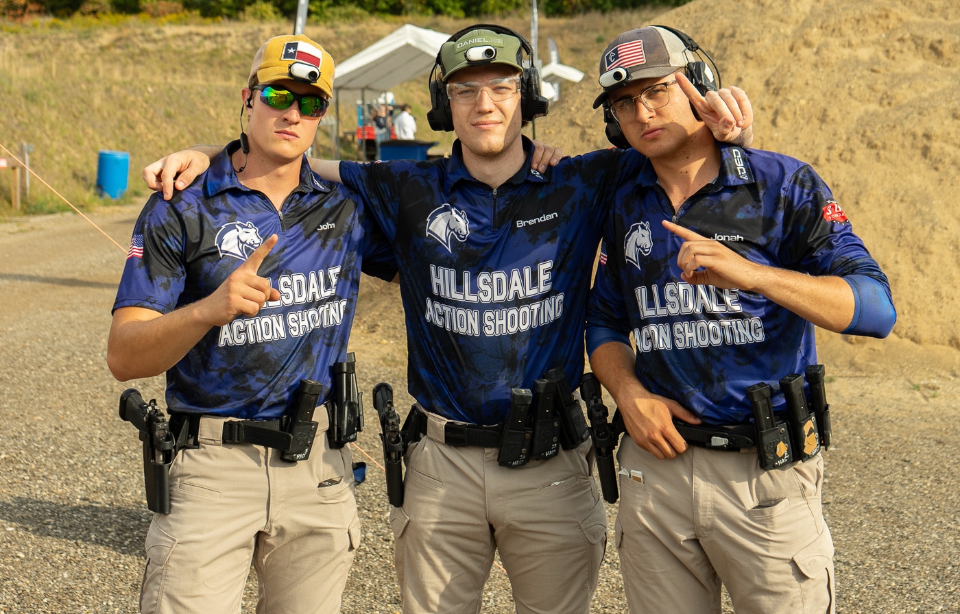 Several Hillsdale Chargers shooting team members stand together during a break in competition at the 2025 USPSA Collegiate Action Pistol Championships held at their Michigan home range. The Hillsdale College competitive shooting athletes relax between stages while waiting for their next runs at the John A. Halter Shooting Center facility. These Hillsdale action pistol team competitors display the team unity and supportive atmosphere that characterizes their marksmanship program. The Hillsdale Chargers pistol shooters include both male and female athletes who competed in Limited and Limited Optics divisions against 13 university teams. Hillsdale College shooting team members like John Beecher, Joseph Grohs, Jonah Kirstein, Brendan Burnham, Kayla Mullin, Clara Bozzay, and Jianna Coppola contributed to second place team finishes. The Hillsdale competitive shooters demonstrate the sportsmanship praised throughout the match where competitors help each other tape targets and encourage newer shooters. These Hillsdale action pistol athletes represent a program that has won back-to-back championships and continues attracting more collegiate teams to compete at the Roger Ailes shooting facility. The Hillsdale marksmanship team members enjoy the beautiful weather conditions that made the 2025 match a success unlike the previous year's competition during Hurricane Helene.