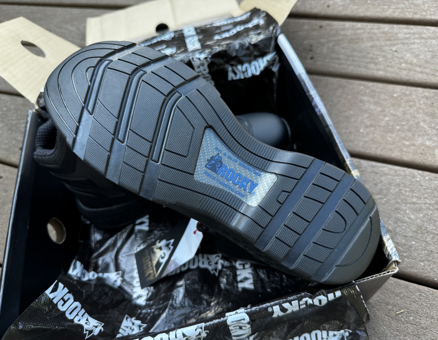 Close-up photograph of Rocky Code Blue Public Service boot non-slip rubber outsole showing oil-resistant slip-resistant tread pattern design for law enforcement tactical footwear. Police duty boot sole displays large lateral grooves and smaller concentric tread patterns preventing hydroplaning on wet pavement surfaces during patrol activities. Rocky Boots six-inch tactical boot bottom features automotive-style tread design without aggressive lugs providing urban patrol traction and professional appearance. Rubber outsole construction of public safety boots shows flat profile suitable for city streets pavement and light terrain navigation for police officers. Non-metallic patrol boot sole attached to breathable mesh upper with TPU overlays demonstrating durable construction and solid bonding between components. Oil-resistant rubber compound on Rocky Code Blue boots provides grip on varied surfaces including wet floors slippery pavement and outdoor terrain encountered during law enforcement duty. Slip-resistant tactical boot outsole tested through field work including drainage ditches overgrown areas and chain-link fence climbing showing versatile traction capability. Budget-friendly police duty boot under $98 MSRP features practical tread pattern balancing slip resistance with ability to maintain professional appearance for public safety officers.