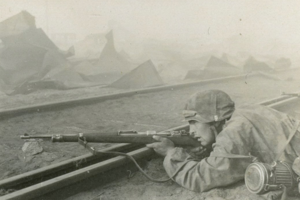 A German sniper takes firing position behind railroad tracks on the Eastern Front, aiming a scope-equipped Karabiner 98k Mauser rifle during World War 2 combat operations against Soviet forces. The Wehrmacht or Waffen SS marksman demonstrates tactical use of railway infrastructure as cover while employing the scoped Kar98k bolt-action sniper rifle that served German designated marksmen throughout the war. This precision shooting variant of the standard German infantry rifle features a telescopic sight mounted to the receiver, transforming the basic Mauser 98k into an effective long-range weapon system. German snipers operated extensively on the Eastern Front from Operation Barbarossa in 1941 through the final battles in Berlin, using the 7.92x57mm chambered Karabiner 98k to engage Soviet infantry, officers, and crew-served weapon teams at extended distances. The railroad tracks provide both hard cover against incoming fire and a stable shooting platform for the German marksman to steady his scoped Mauser rifle for precision shots. Wehrmacht sniper doctrine emphasized two-man teams and aggressive forward positioning, with SS units particularly known for using stay-behind sniper elements to harass advancing Allied and Soviet troops during tactical withdrawals. The Kar98k sniper rifle could be fitted with various German military optics including the 4X ZF4 scope, the turret-mounted Zeiss ZF39, or the less popular 1.5X ZF41 long eye relief sight. German snipers on the Eastern Front became legendary for their effectiveness, with the scoped Karabiner 98 kurz serving as their primary weapon throughout the brutal campaign against the Red Army.