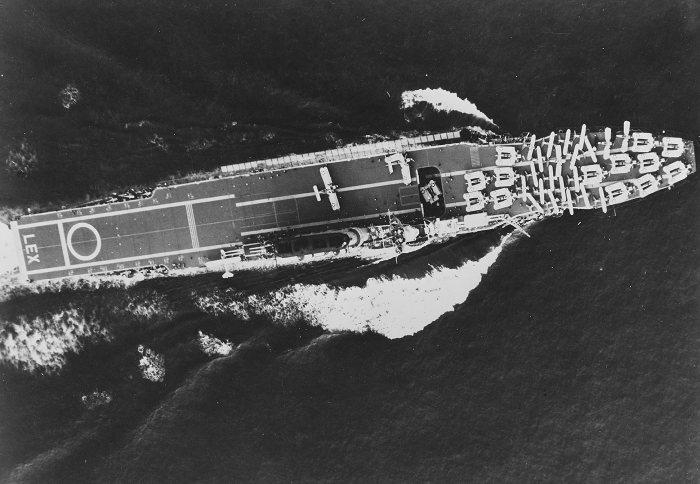 Overhead photograph of USS Lexington CV-2 underway in March 1932 showing the massive 901-foot flight deck of the converted Lexington-class battlecruiser carrier with aircraft arranged on deck. Lady Lex operated with 86 warplanes during this peacetime period when the United States Navy was developing and refining carrier aviation tactics through extensive naval exercises. USS Lexington aircraft carrier had participated in Grand Joint Exercise No. 4 just one month earlier in February 1932, conducting a simulated surprise attack on Pearl Harbor. The Lexington CV-2 was commissioned in late 1927 under the Washington Naval Treaty that allowed battlecruiser hulls to be converted into carriers with 36,000-ton displacement. This overhead view shows the flight deck that was 100 feet wide with innovative lowerable crash barriers enabling the carrier to quadruple aircraft landing rates compared to other flattops. USS Lexington and sister ship Saratoga CV-3 were the largest aircraft carriers in the world operating twice the number of warplanes as comparable Royal Navy carriers. Lady Lex was still considered experimental when this photograph was taken but would prove highly successful during World War II Pacific operations before being lost at the Battle of Coral Sea in May 1942.