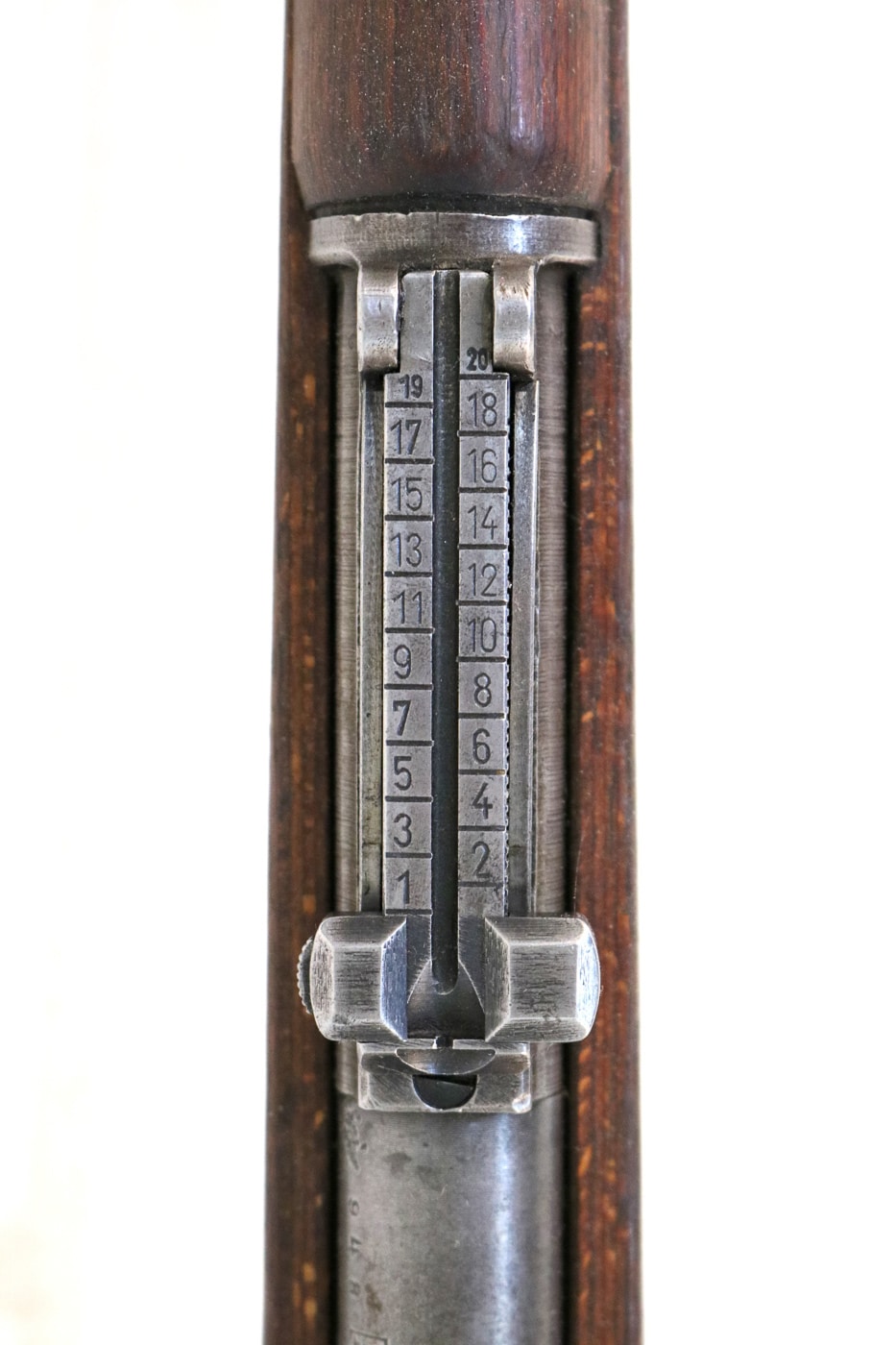 Close-up photograph of the Karabiner 98k sliding tangent leaf rear sight showing the graduated markings calibrated in 100-meter increments from 100 out to 2,000 meters for long-range shooting with the German bolt-action rifle during World War 2. The Kar98k rear sight assembly mounts on the barrel just forward of the receiver, featuring a folding tangent leaf that German soldiers adjusted up or down to compensate for bullet drop at different ranges when firing the 7.92x57mm Mauser cartridge. The graduated sight markings allowed Wehrmacht infantry to dial in specific range settings from close quarters out to theoretical maximum effective distance, though practical combat accuracy with iron sights rarely exceeded 400 to 600 meters even for skilled marksmen. German military doctrine emphasized marksmanship training with the Mauser 98k, with soldiers learning to estimate range and adjust the tangent rear sight accordingly during training exercises before deployment to combat zones. The sliding sight leaf on the Karabiner 98 kurz pairs with a fixed barleycorn front sight to create the traditional iron sight system used on military bolt-action rifles throughout the early 20th century. Rifles manufactured after 1939 incorporated a pressed steel hood over the front sight to protect it from damage during combat, transportation, and field handling that could knock the sight out of alignment. The 2,000-meter maximum graduation on the Kar98k rear sight reflects optimistic expectations for infantry rifle effectiveness, though actual combat engagements typically occurred at much shorter ranges where the tangent sight settings of 200 to 400 meters saw more practical use. German soldiers learned to quickly adjust the sliding rear sight during combat based on target distance, with experienced infantry developing instinctive understanding of proper sight settings for common engagement ranges encountered on the Eastern Front, Western Front, and other theaters throughout World War 2.