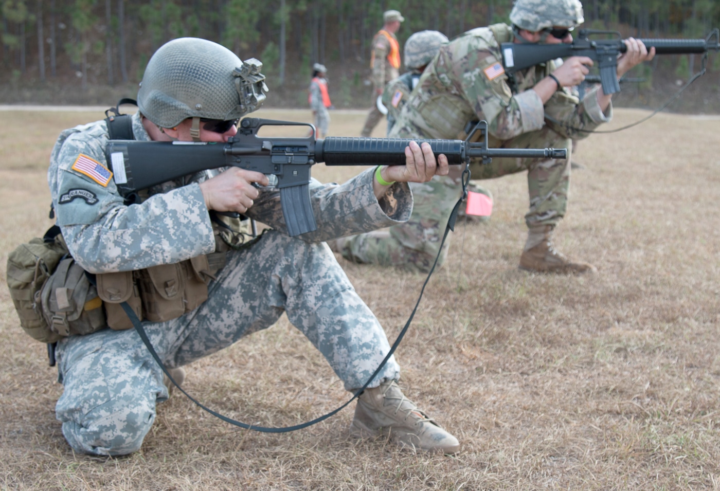 Military service member fires M16A2 rifle during marksmanship competition demonstrating accuracy improvements over M16A1 variant design. The M16A2 features fully adjustable rear sight with click adjustments for windage and elevation from 300 to 800 meters compared to basic flip aperture on M16A1. Flat-faced front sight post on A2 with four elevation positions eliminates optical aberrations caused by round post on M16A1 in certain lighting conditions. Bird cage flash hider on M16A2 with bottom slots omitted reduces muzzle rise during recoil unlike three-prong duck bill flash suppressor on A1 variant. Heavier barrel profile forward of handguard on A2 provides better accuracy and heat dissipation than thinner M16A1 barrel configuration. Barrel twist rate of 1:7 inches on M16A2 stabilizes heavier 62-grain M855 ammunition better than 1:12 twist on M16A1 designed for 52-grain M193 rounds. Extended length of pull stock on A2 with textured buttplate improves shooting stability compared to shorter smooth stock on M16A1. Competition scenario illustrates M16A2 precision shooting capabilities and long-range accuracy enhancements requested by Marine Corps after Vietnam War experience with M16A1 performance at 25-meter engagement distances versus modern battlefield requirements at 300 to 400 meters.
