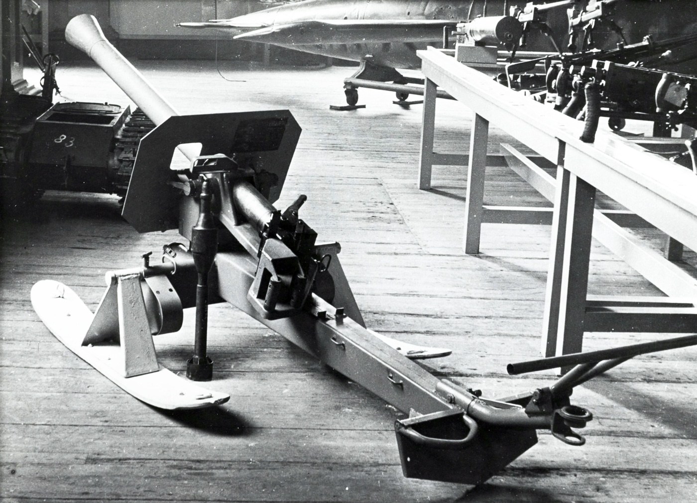 A preserved 8.8cm Raketenwerfer 43 Püppchen anti-tank rocket launcher sits on display in a military museum, configured with wooden ski mounts instead of its standard wheeled carriage. Museums preserve German World War II weapons. The Püppchen on skis shows winter configuration. Military collections display Raketenwerfer 43 examples. The ski-mounted launcher has an extremely low profile. German anti-tank weapons featured interchangeable mounts. Museum visitors can examine the Püppchen design. The 88mm rocket launcher survives in collections. Historical displays show Wehrmacht equipment variations.